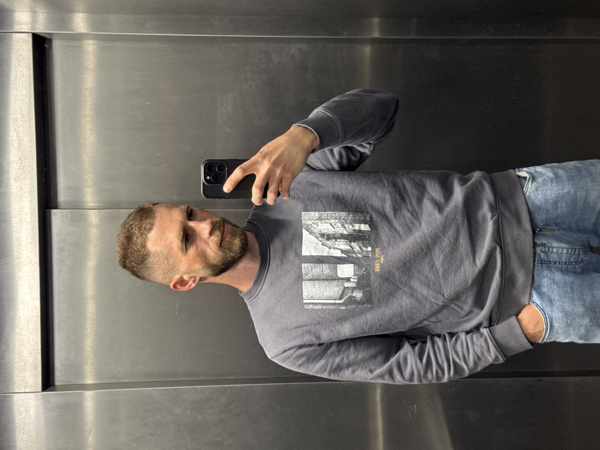 A man with short blonde hair and a beard is taking a selfie in what appears to be an elevator with a stainless steel interior. He is wearing a grey sweatshirt with a black and white graphic on the front and blue jeans. He is holding a black smartphone in his right hand, which is positioned to capture his reflection. The image is slightly rotated counter-clockwise. Based on the "PEPE JEANS" text on the sweatshirt, which is reflected backwards, the scene is likely unrelated to the city of Budva, Montenegro.