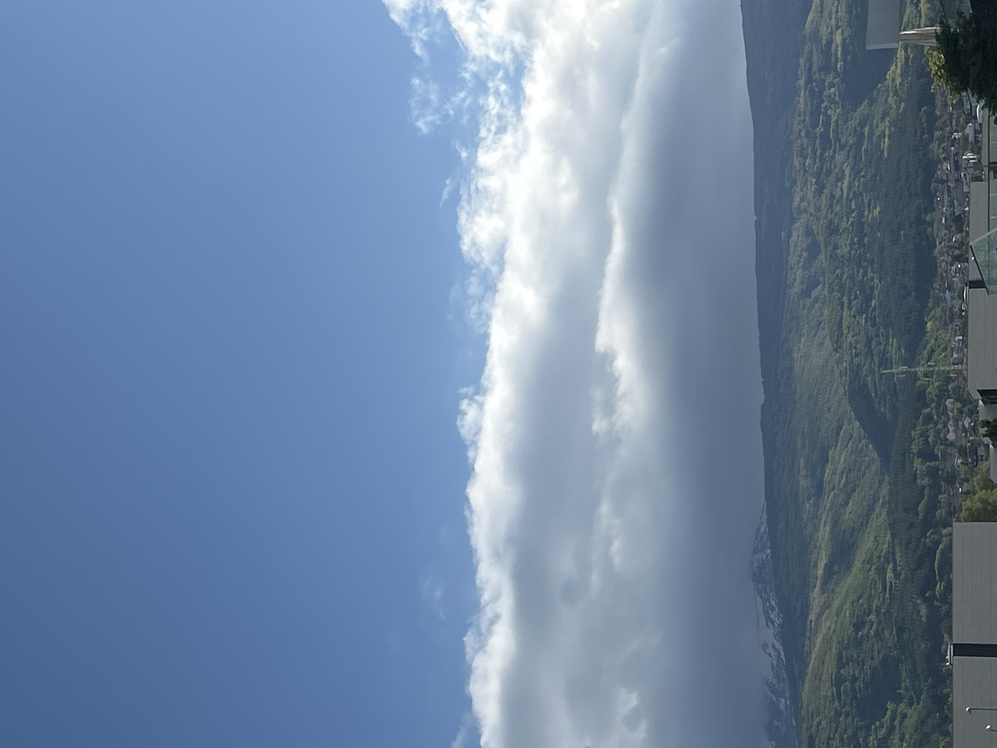 The image shows a landscape with a clear blue sky on the left side and a large bank of clouds in the middle, next to a mountain covered in green vegetation on the right side. Buildings and structures are visible at the foot of the mountain. I don't know the location.