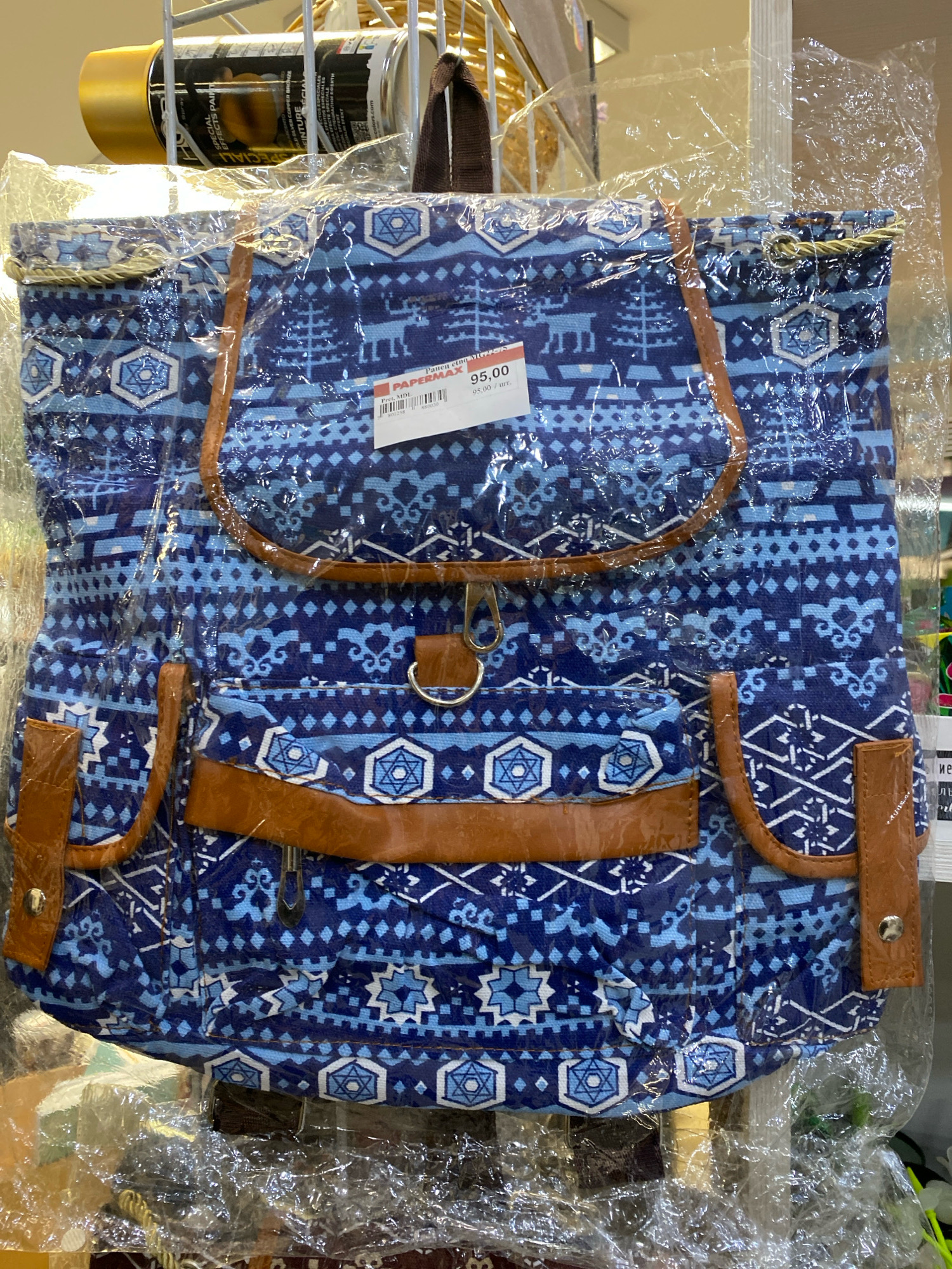 A blue fabric backpack with a white and light blue geometric pattern, featuring images of reindeer and trees, is displayed in a retail setting. The backpack is enclosed in clear plastic packaging and has brown faux-leather trim around the pockets and top flap. A price tag reading "95,00" is attached to the front of the backpack. The backpack hangs from a metal rack, and in the background, other items, including a can of gold spray paint, are visible. The overall environment suggests a shop, likely in Chișinău, Moldova, given the price format and the currency. There are no people visible in the image.