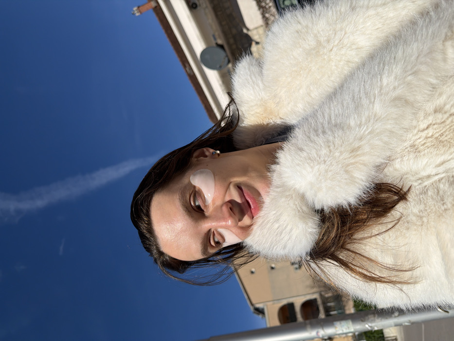 The image displays an individual, identified as female, from the upper chest upwards, positioned outdoors in an urban environment in Budva, Montenegro. The subject has long, dark brown hair and a smiling facial expression, with her gaze directed slightly towards the viewer's upper left. Two translucent, heart-shaped cosmetic patches are applied beneath each eye. A small, silver-colored stud earring is visible in her right ear. She is attired in a voluminous, off-white fur or faux fur coat featuring a prominent collar. The background consists of a clear, vivid blue sky with a single white aircraft contrail extending diagonally from the upper left. To the far left, a metallic pole is partially visible. Behind the subject, architectural elements include a light-brown building with arched window structures on the left, and a light-colored building with a reddish-brown tiled roof and a dark grey satellite dish mounted on its wall on the right. The scene is brightly illuminated by direct sunlight.