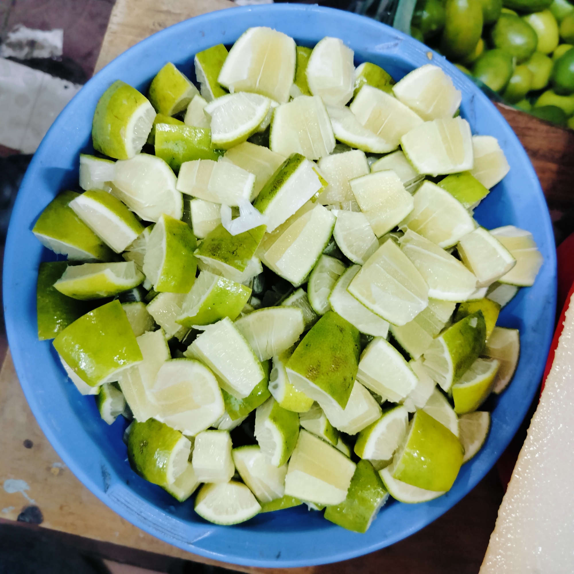 In a close-up, overhead shot, a bright blue plastic bowl is filled with chopped lime wedges. The limes are cut into roughly equal, bite-sized pieces, showcasing both their vibrant green rind and pale yellow-white flesh. Some pieces have a slightly translucent quality, suggesting they are juicy. The bowl sits on a wooden surface, which appears to be a tabletop or market stall. In the blurred background, more whole green limes are visible, along with other indistinct market items. The overall impression is of fresh produce ready for preparation, possibly for a drink or dish. The setting suggests a market in Dhaka, Bangladesh, where limes are a common ingredient and are often sold in bulk.