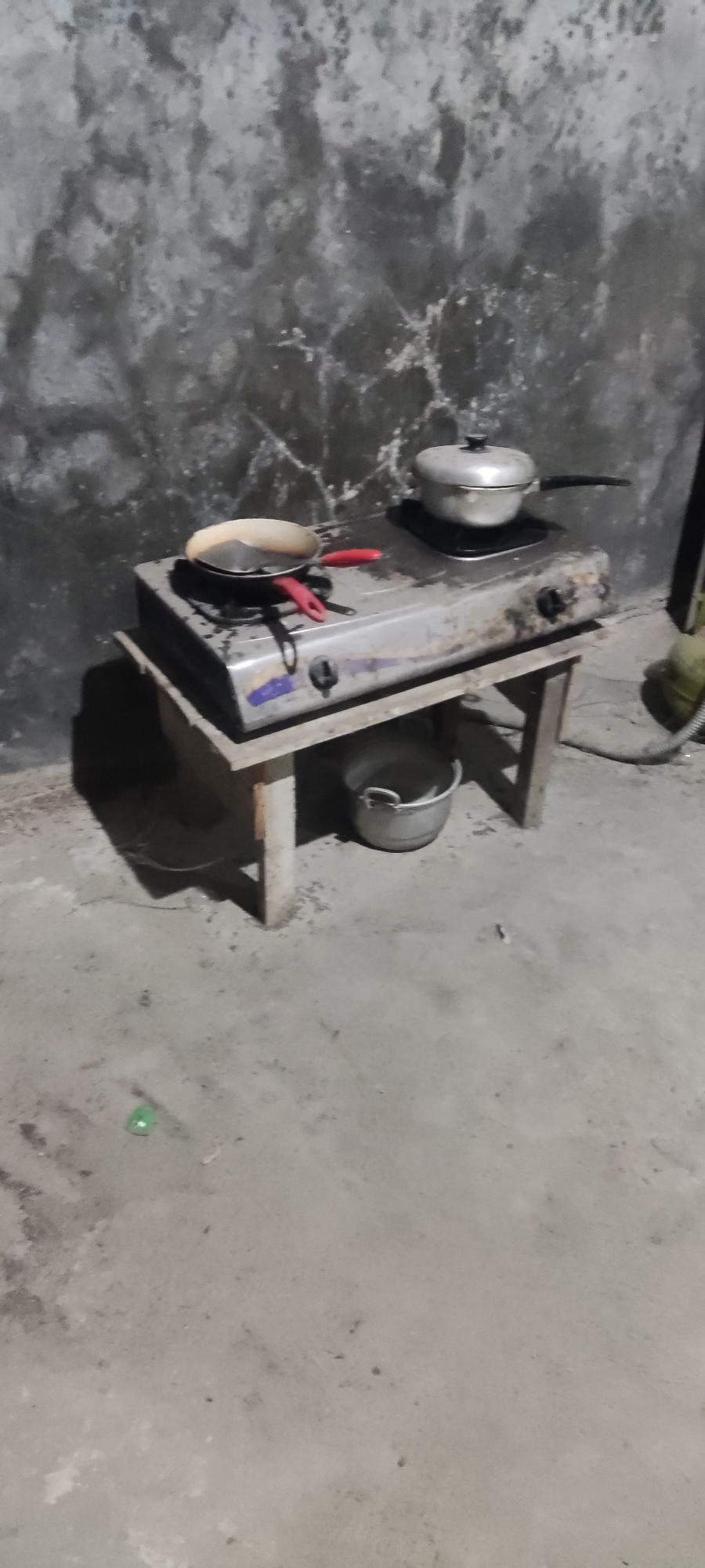 The image depicts an outdoor cooking scene, possibly in Karangwangun, Indonesia. In the foreground, a weathered concrete floor is visible, with some debris scattered across it. A simple wooden table supports a metal two-burner gas stove. On the stove, a frying pan with a red handle contains some food remnants, and a pot with a lid is also present. Beneath the table, a metal bucket sits on the ground. The background features a textured, rough wall, likely made of concrete or plaster, exhibiting signs of age and weathering. The lighting suggests it may be evening or a dimly lit area. The scene appears utilitarian, suggesting a functional cooking setup rather than a formal kitchen environment.