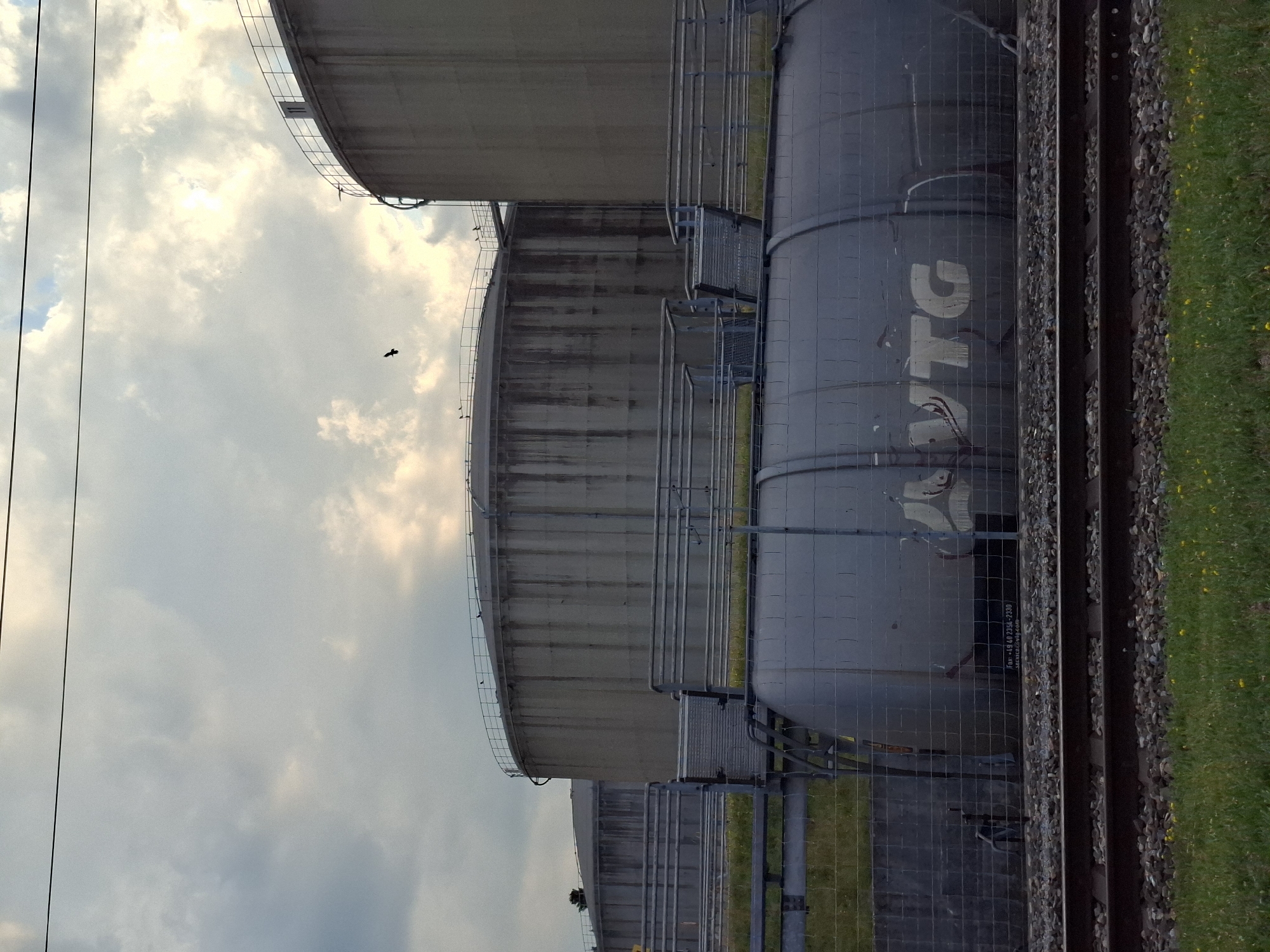 The image shows a large industrial area with several cylindrical storage tanks and a train car. The storage tanks are made of concrete and have a weathered appearance. One tank in the foreground is a dark color and has the "VTG" logo visible on its side. The train car is also dark and appears to be a tanker car. There are train tracks in front of the train car. In the background, there are more storage tanks, some of which are taller than the one in the foreground. The sky is cloudy with patches of sunlight breaking through. There is a single bird in flight in the upper portion of the image. The scene appears to be an industrial facility located near Rothenburg, Switzerland.