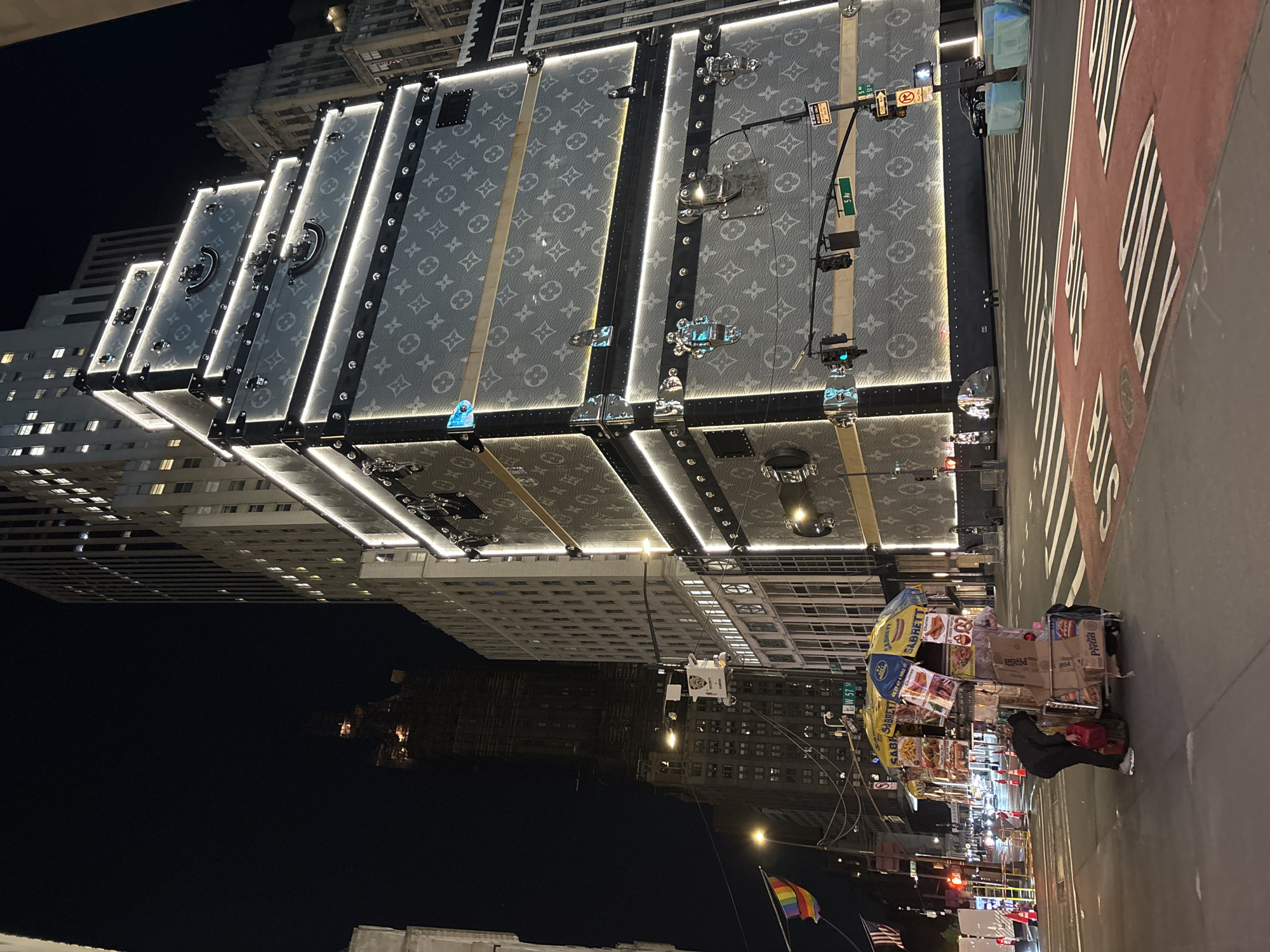 The image captures a vibrant night scene on a bustling street in New York City. The prominent subject is a tall building whose facade has been dramatically transformed to resemble several colossal, stacked Louis Vuitton trunks. These oversized trunks feature the distinctive grey Louis Vuitton monogram pattern and are brightly illuminated along their edges and straps with glowing LED lights, creating a striking visual spectacle against the dark sky. The building behind this display shows numerous lit windows, indicative of an urban skyscraper.
At street level, the scene depicts typical city life at night. A street vendor operates a hot dog cart, identifiable by its yellow and blue "Sabrett" umbrella, offering snacks and drinks. A person is partially visible near the cart. The street itself has "BUS" painted markings and is lined with traffic lights and street signs. Several signs clearly indicate the location, displaying "5 Av" (5th Avenue) and "W 57 St" (West 57th Street), along with regulatory signs like "ONE WAY" and "NO TURN ON RED." A rainbow Pride flag and an American flag are visible at the bottom of the frame, adding to the urban tapestry. The overall atmosphere is one of a busy, illuminated metropolis after dark, with various city lights contributing to the dynamic environment.