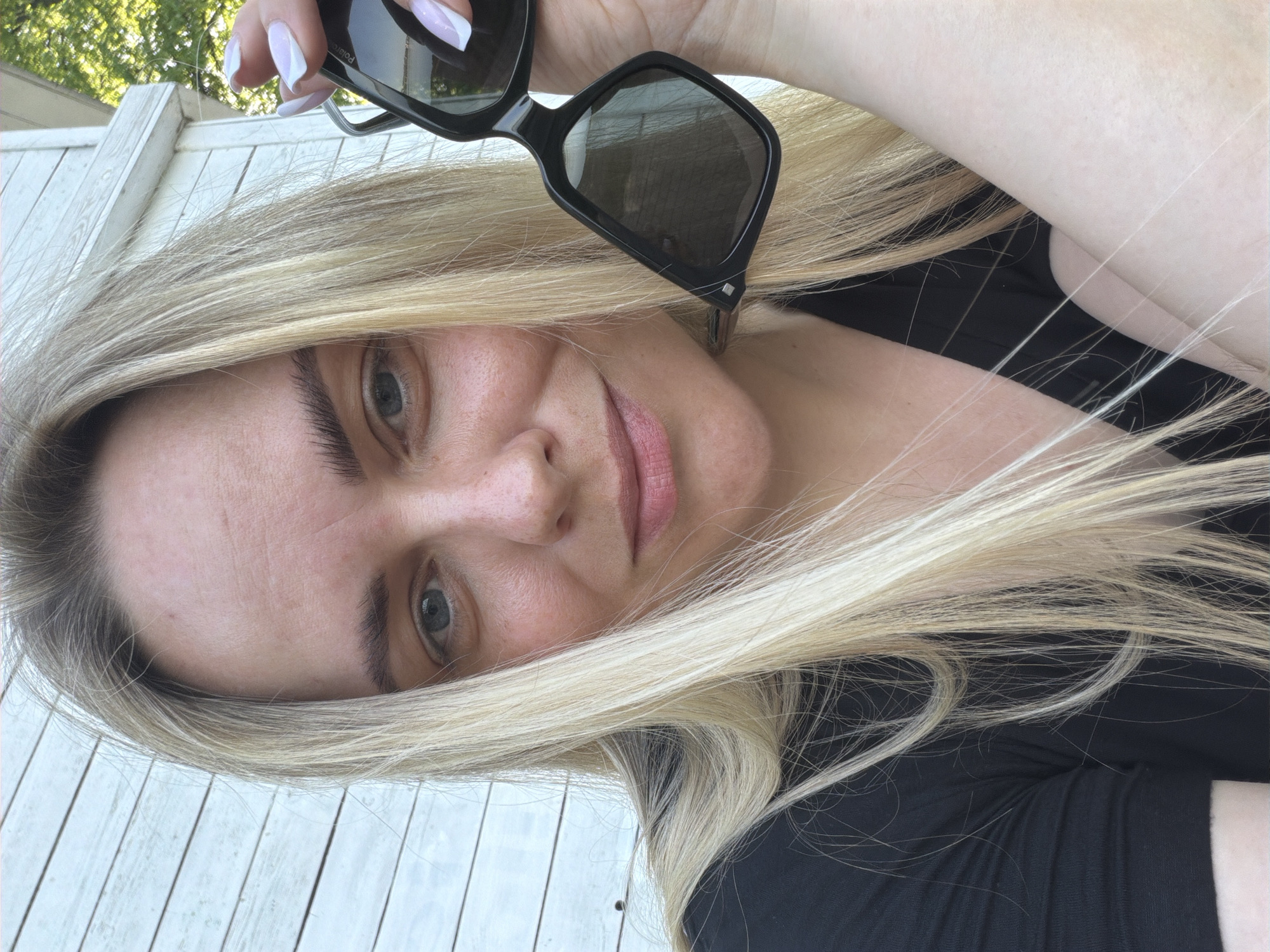 A person with long blonde hair and fair skin is holding a pair of black sunglasses up near their face. They are wearing a black shirt. The background includes a white, slatted wall and green foliage. The location is not identifiable as being in Votkinsk, Russia based on the provided image.