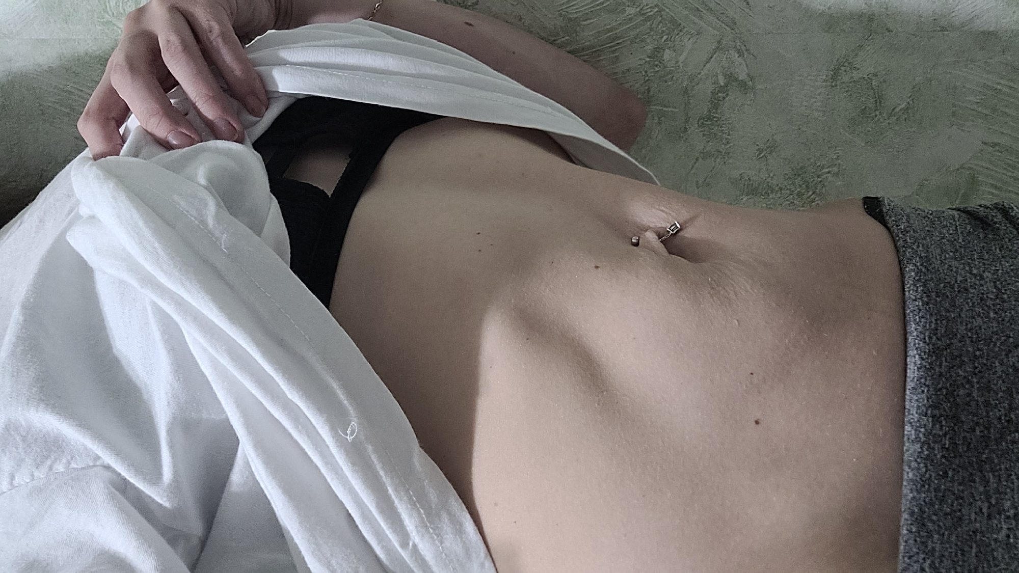 A person's torso is shown, with a white shirt lifted to reveal the abdomen. The individual has a navel piercing. The person is lying down, and the background is a textured wall.