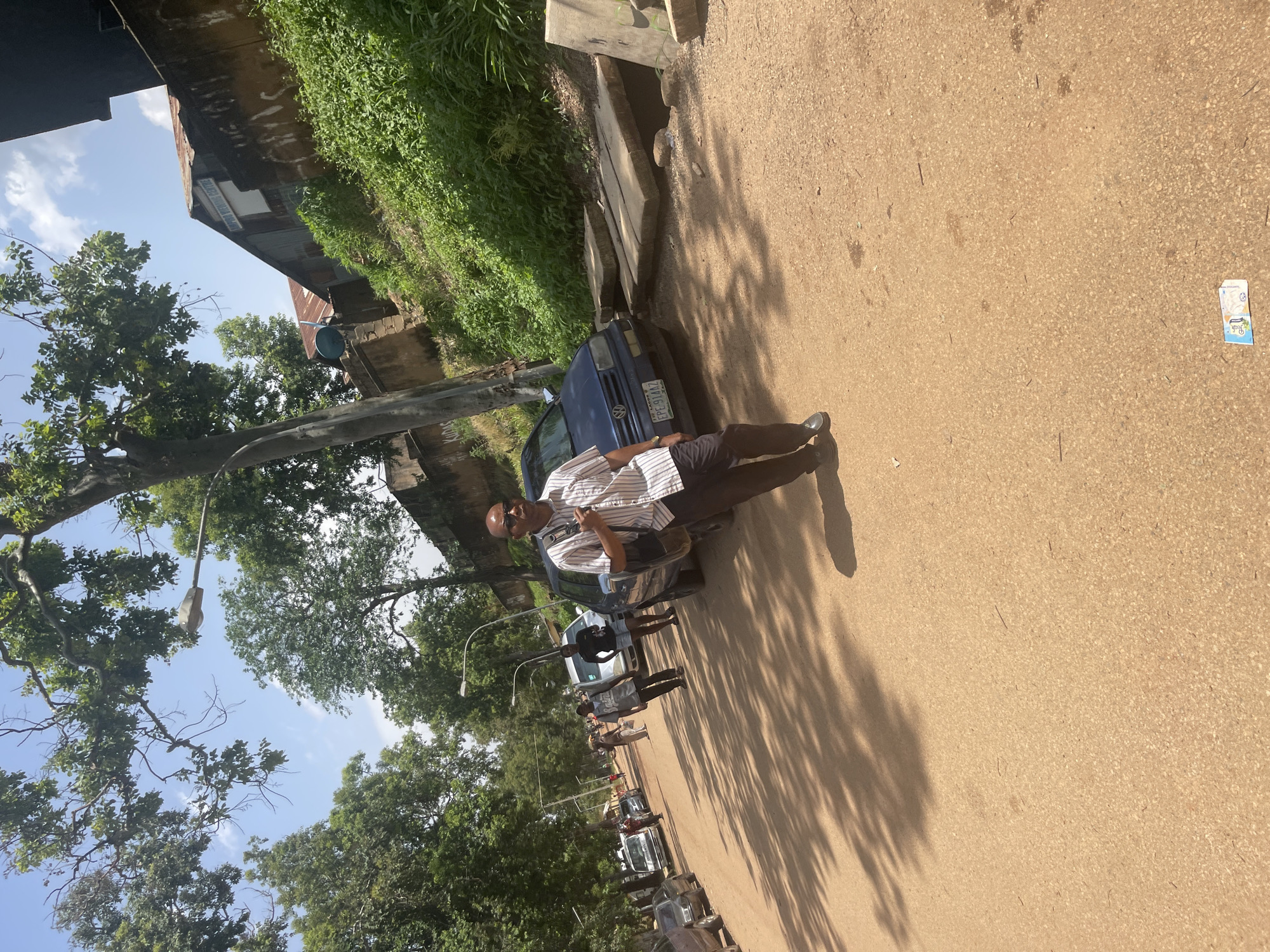 The image depicts a street scene in Rafin Par, Nigeria. A man in a striped shirt and dark pants walks towards the camera on a sandy road. He is wearing sunglasses and carrying a bag over his shoulder. Several cars are parked along the road, including a blue Volkswagen. In the background, there are trees, buildings, and a lush green hillside. A street lamp is visible on the right side of the road. The sky is bright blue with some clouds. The overall impression is a sunny day in a semi-urban environment.
