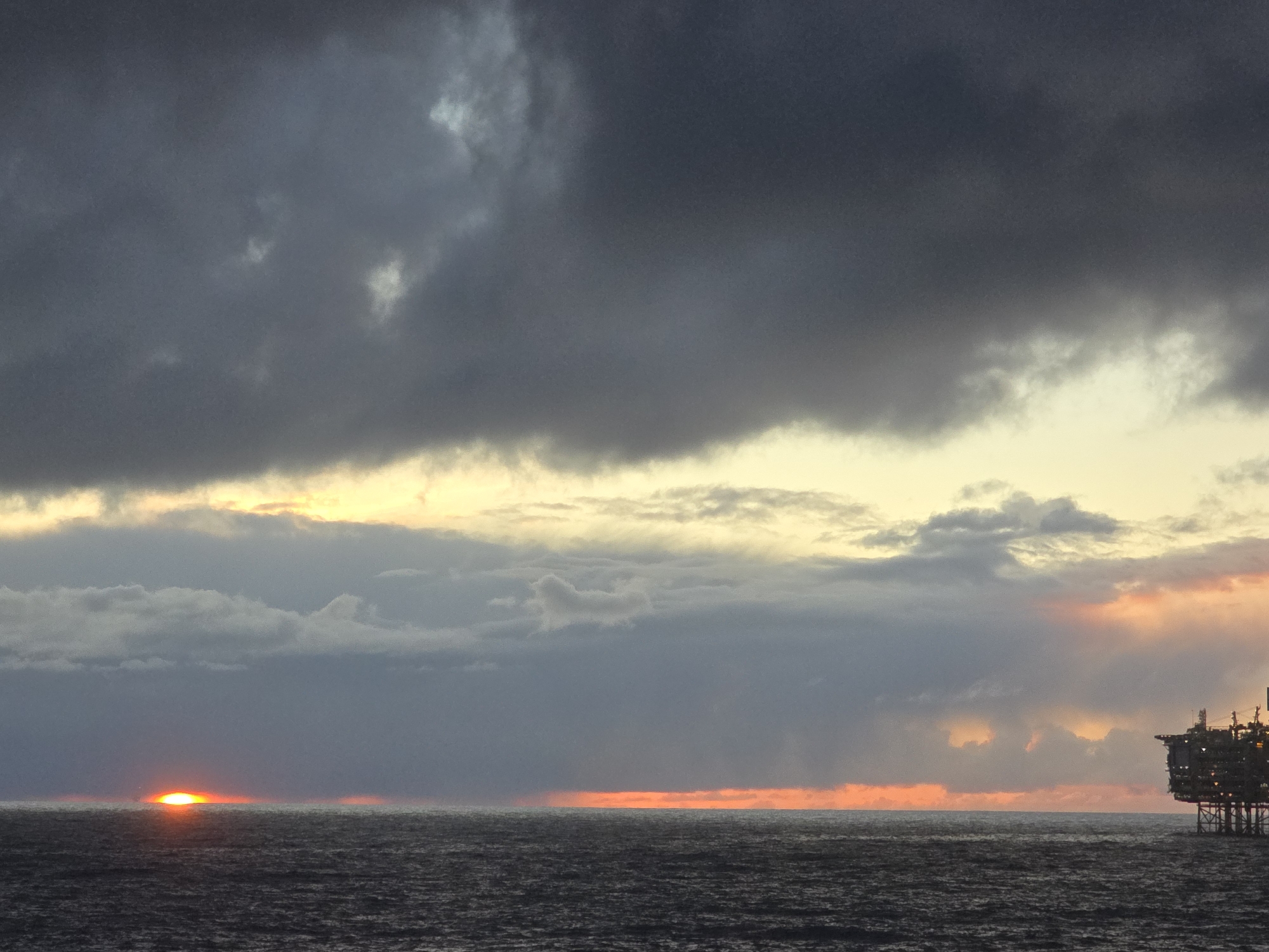 The image depicts a seascape at either sunrise or sunset, characterized by a low horizon line and predominant cloud cover. The sky is dominated by dark, ominous clouds in the upper portion, transitioning to lighter, more diffused cloud formations closer to the horizon. The sun, or its afterglow, appears as a fiery orange streak along the horizon, reflecting faintly on the water's surface. An offshore oil platform is visible on the right side of the frame. The sea itself appears dark and choppy.
Location: Unknown, Unknown.