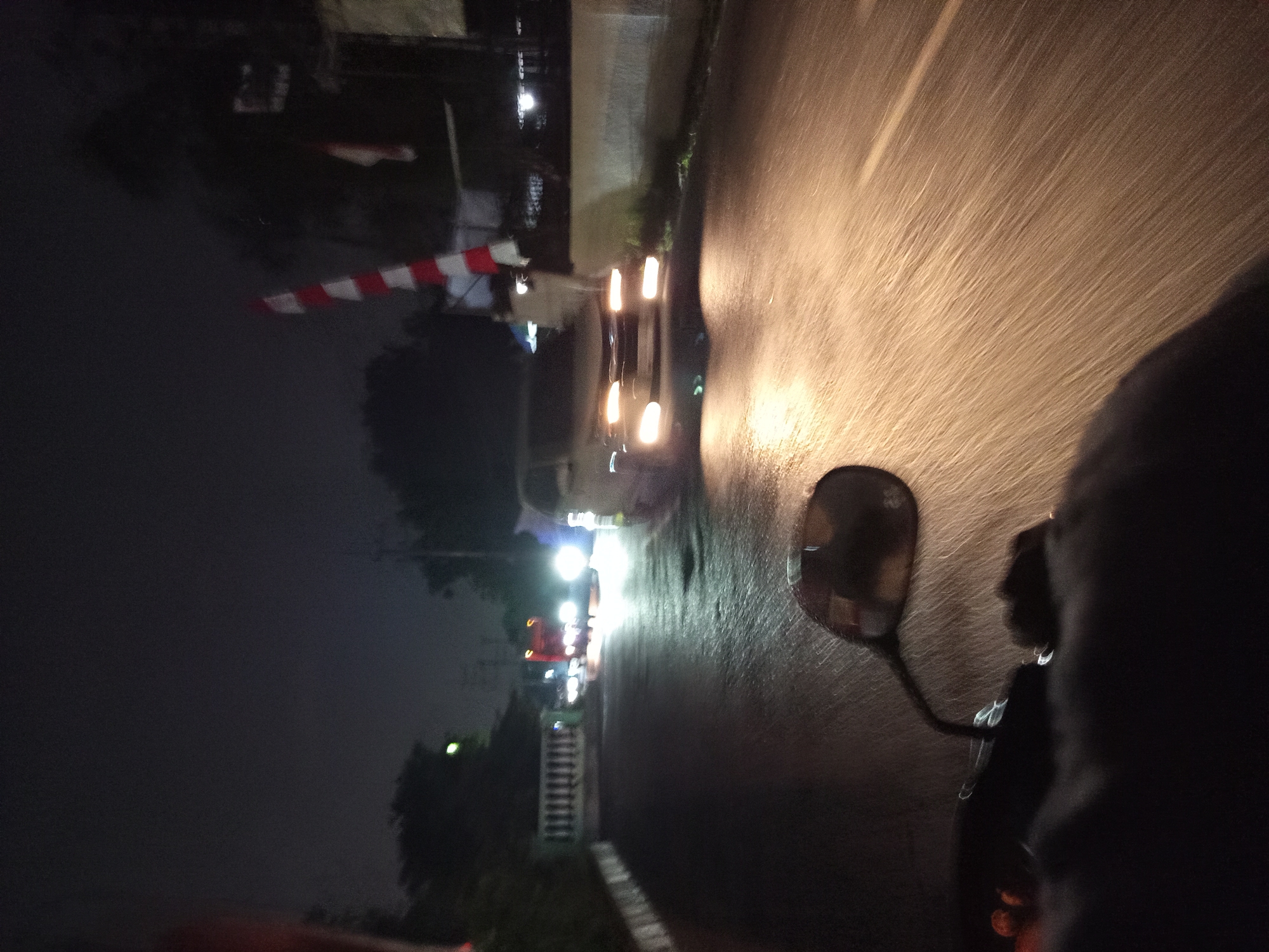This image depicts a nighttime scene on a wet road, likely in Sruni, Indonesia, characterized by blurred motion. The perspective appears to be from a motorcycle, with a dark silhouette of the rider and a portion of the handlebar and mirror visible in the foreground. The road surface reflects the bright headlights of oncoming traffic, creating streaks of light that emphasize the movement.
In the middle ground, a white car with illuminated headlights is in motion, positioned on the right side of the road. Further down the road, a red truck with its hazard lights on is also visible. To the right of the road, a red and white striped barrier is partially visible, suggesting a checkpoint or crossing. Trees and foliage line the sides of the road, partially obscuring structures in the background. The overall atmosphere is dark, with artificial lights illuminating the immediate vicinity and creating a sense of speed and transient activity.