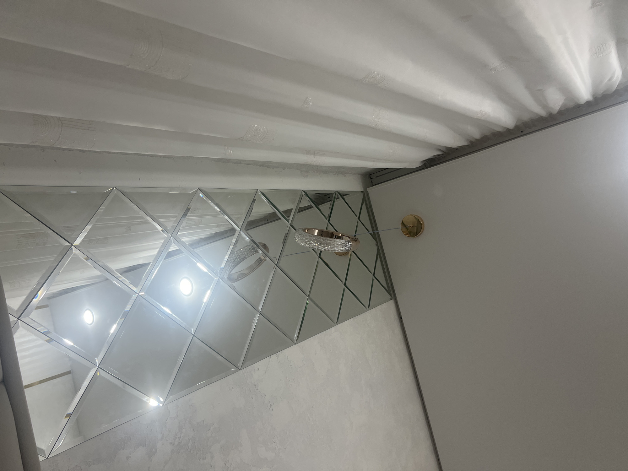 In a room, white sheer curtains hang to the left. A wall to the right is covered in diamond-shaped mirrors. The ceiling is white. A gold light fixture with a crystal pendant hangs from the ceiling above the mirrored wall. Two lights are visible reflected in the mirrors. The image appears to be an interior shot, likely a living room or dining area in Veteran-2, Russia.
