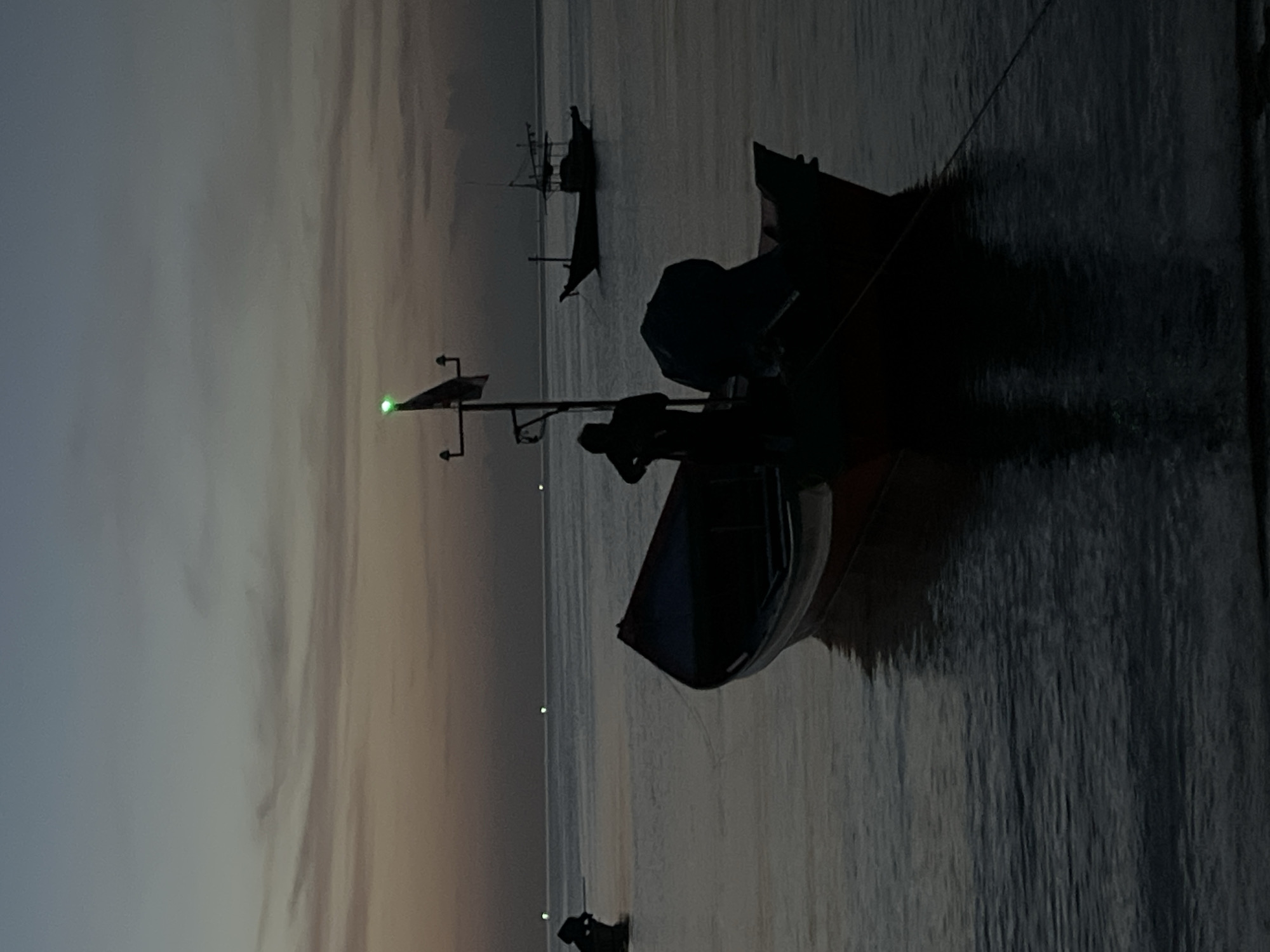 The image depicts a serene evening scene on the coast of Ko Pha-ngan District, Thailand. The sky, transitioning from dusk to night, displays muted hues of blue, gray, and orange, with thin, wispy clouds. The calm sea stretches towards the horizon, punctuated by faint, distant lights. In the foreground, a dark silhouette of a fishing boat is anchored, its outline softened by the dim light. A person stands on the boat, their form indistinct. Another similar boat is visible further out at sea, also in silhouette. The water reflects the subdued light, creating a tranquil and slightly mysterious atmosphere. The overall impression is one of quiet solitude and the peaceful conclusion of a day at sea.