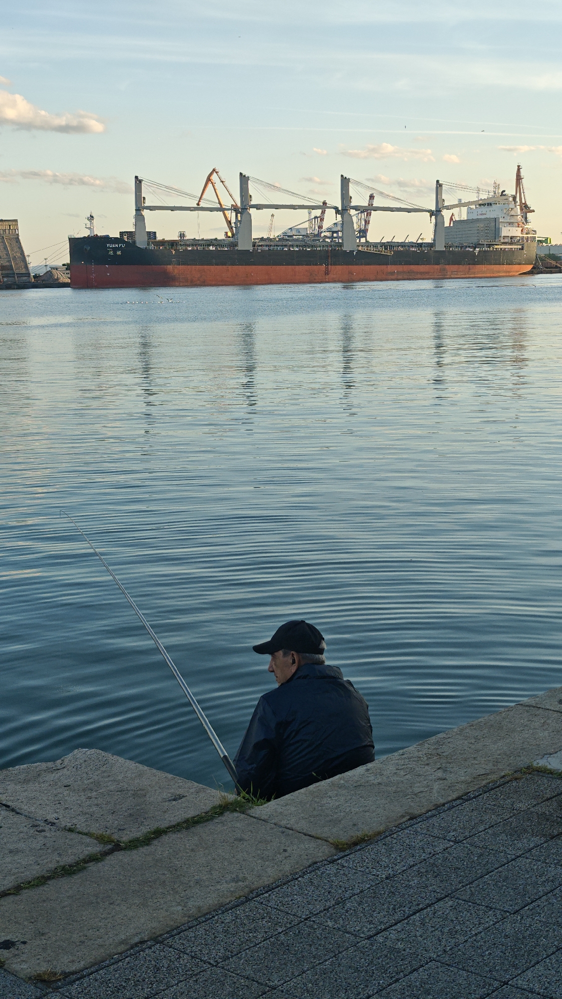 A long shot depicts a man fishing along the Burgas embankment with a large cargo ship in the background, on a clear, sunny day.
The man, wearing a dark jacket and a baseball cap, sits on a concrete edge, holding a fishing rod that extends into the water. The water is calm with subtle ripples.
In the background, a sizable cargo ship, painted with a black and red hull, is anchored, reflected in the water. Cranes and other port infrastructure are visible behind the ship. The sky above is mostly clear, with a few scattered clouds.
The location is the Burgas embankment, Burgas, Bulgaria.