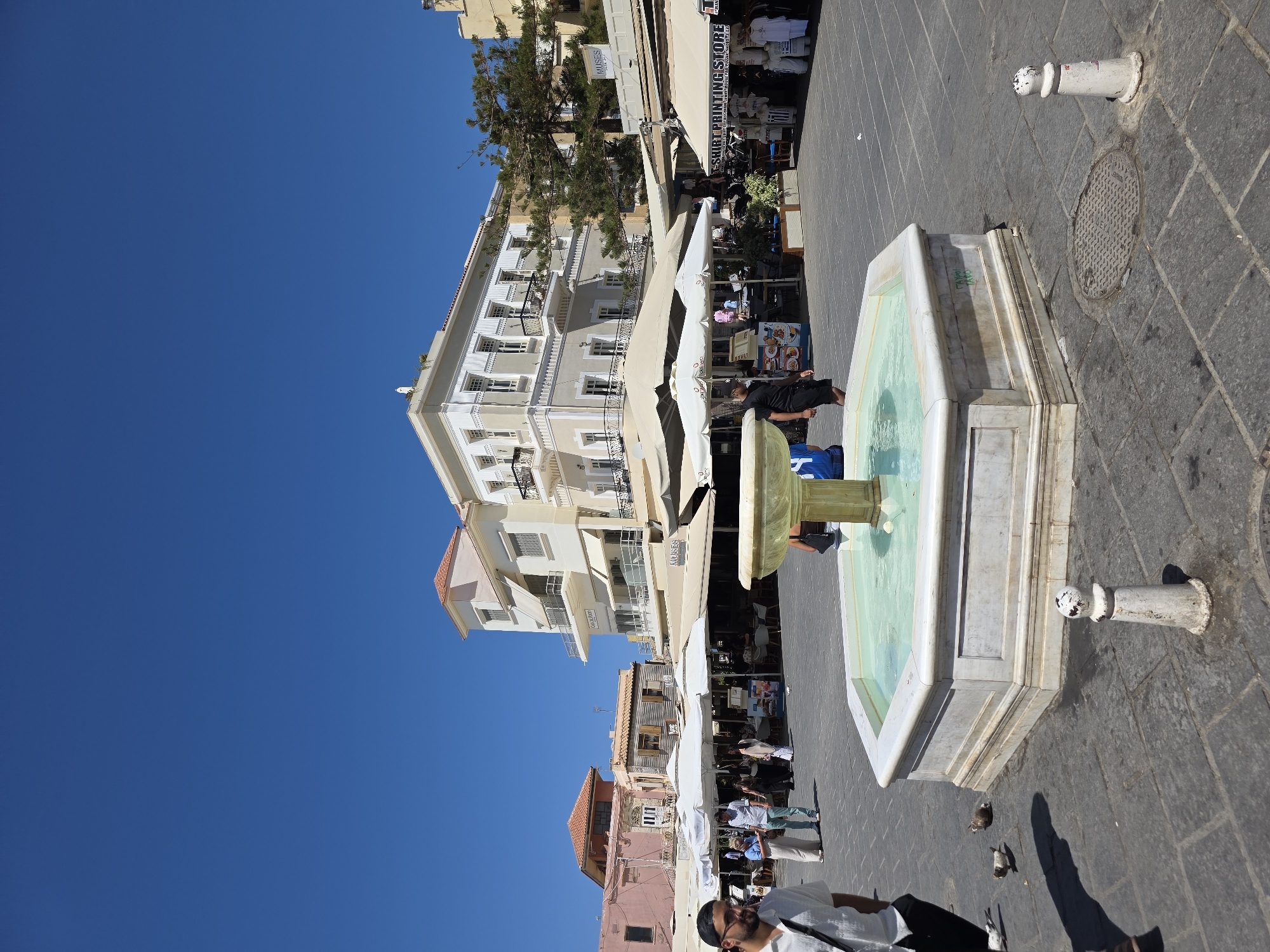 The image captures a sunny day in Chania, Greece, likely in the Old Town, given the architectural style. In the foreground, an octagonal marble fountain with turquoise water is visible, with its base resting on a cobblestone square. Several pigeons are scattered on the ground around the fountain, with one in flight and two pecking at something.
To the left of the fountain, a man wearing a white short-sleeved shirt and dark pants is walking, with a black bag slung over his shoulder. His left hand is in his pocket.
In the background, there are several buildings with traditional European architecture, featuring balconies and awnings. Some of these buildings house shops and cafes, indicated by signs such as "Muses" and "Shirt Printing Store". White umbrellas are set up outside some of the establishments, suggesting outdoor seating. People are seen walking around, sitting at tables, and browsing the shops. The sky is a clear, bright blue. The overall atmosphere is one of a lively public square.