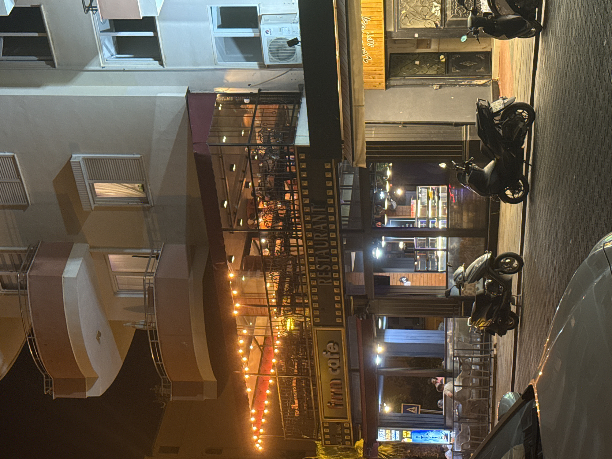 The image depicts a nighttime street scene in Alanya, Turkey, focusing on the facade of a building housing a cafe and restaurant. A multi-story building with balconies overlooks the street. The ground floor features "firm cafe" and "restaurant" signage illuminated by string lights and interior lighting. Tables and chairs are visible both on a covered patio area and inside. Two motorcycles are parked on the cobblestone sidewalk in front of the establishment. A white car is partially visible in the foreground. The lighting suggests it is evening or night.