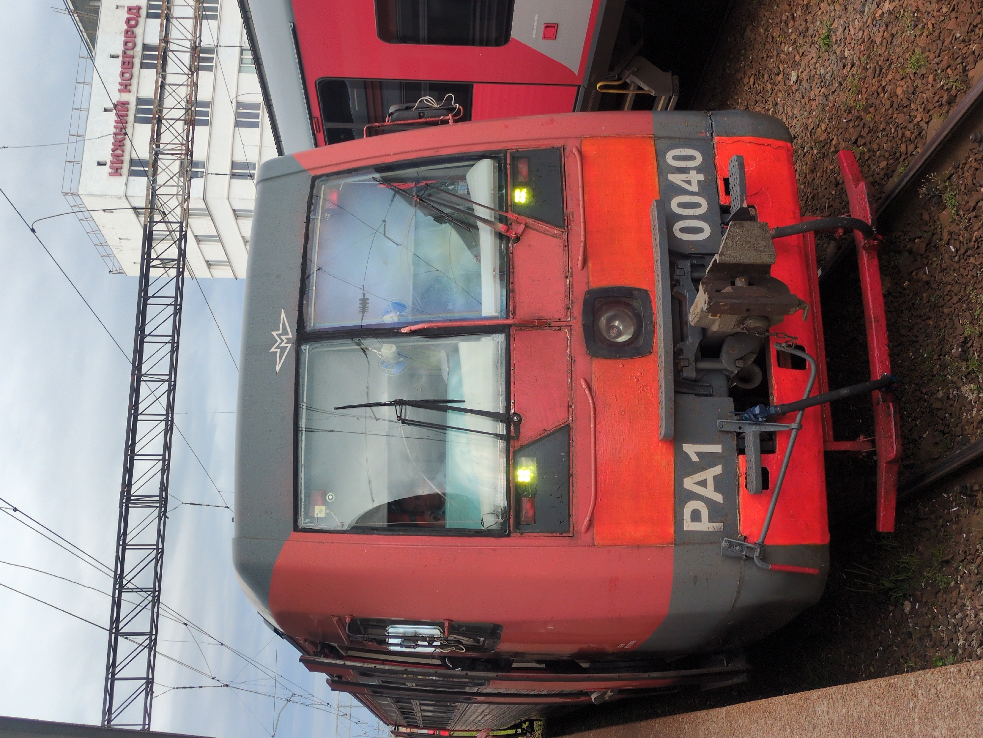 A frontal view of a red and gray electric train is captured at a train station in Nizhny Novgorod, Russia. The train, identified by the markings "PA1" and "0040" on its front, is parked next to a platform. Above the train, overhead wires and their supporting structure are visible against a cloudy sky. In the background, a white building with the text "НИЖНИЙ НОВГОРОД" (Nizhny Novgorod) in red letters is prominently displayed. The train features a large windshield with wipers and two headlights. The surrounding area consists of gravel and train tracks. There are no visible people in the image.