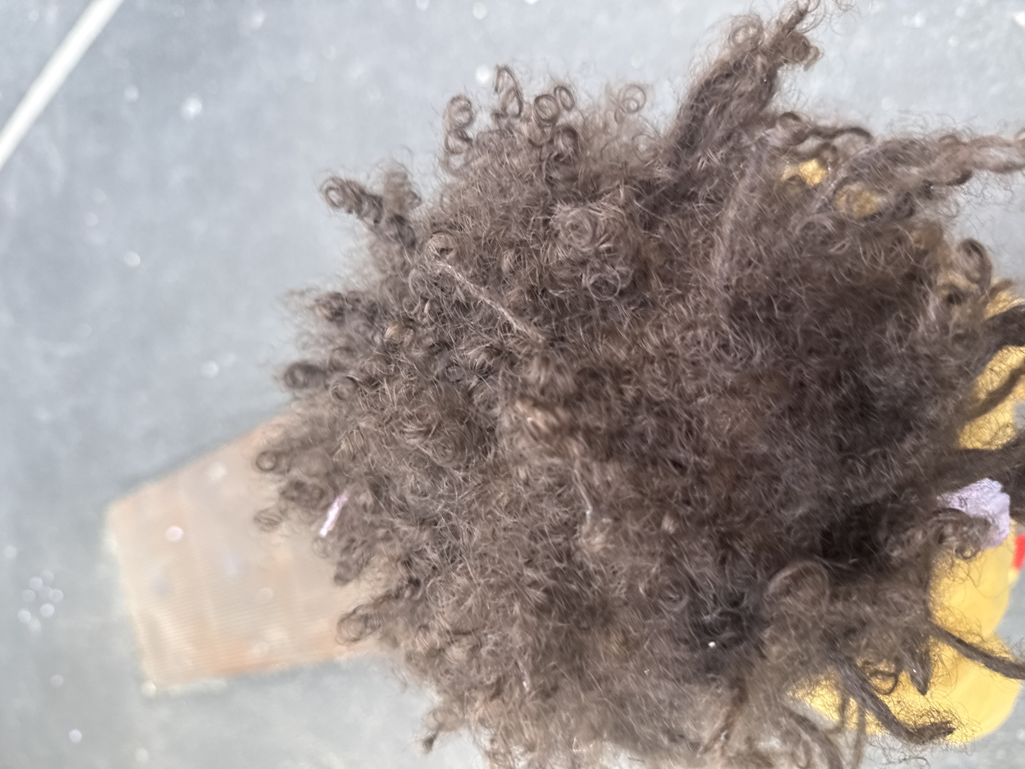 A close-up, overhead shot shows a dense mass of dark, tightly coiled hair, likely from a doll or wig. The hair appears to be situated above a yellow container or object and a clear plastic container. The background has a gray, slightly reflective surface.
The location is not discernible from the image.