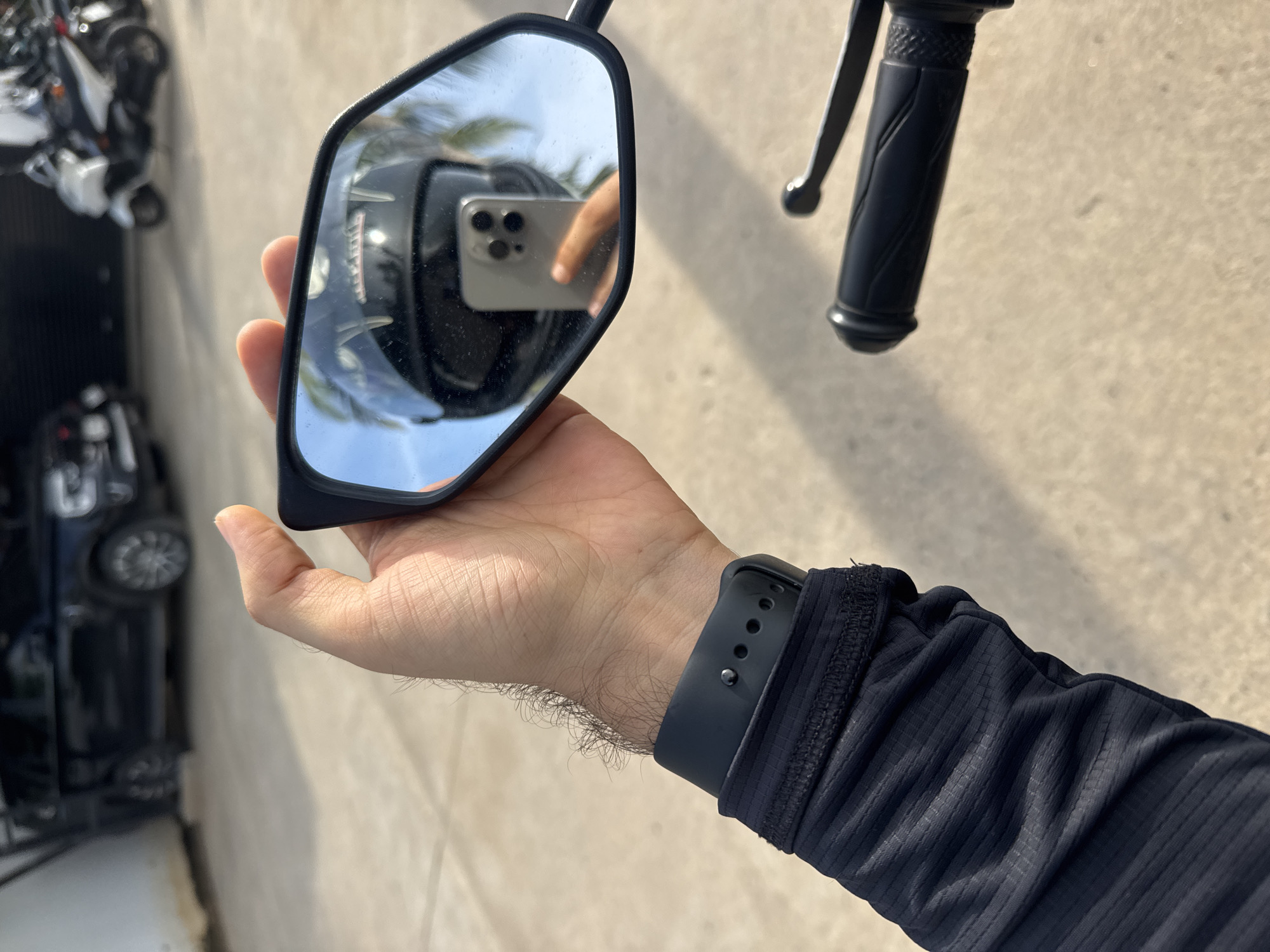 A person's hand, wearing a black smart watch and a black arm sleeve, holds a motorcycle's side mirror. Reflected in the mirror is a black motorcycle helmet and the person's other hand holding a smartphone. In the background, a black SUV and several white scooters are parked on a paved surface. The scene appears to be outdoors, possibly in a parking area, under a bright sky with palm trees visible in the reflection. The location is identified as Ko Samui District, Thailand.