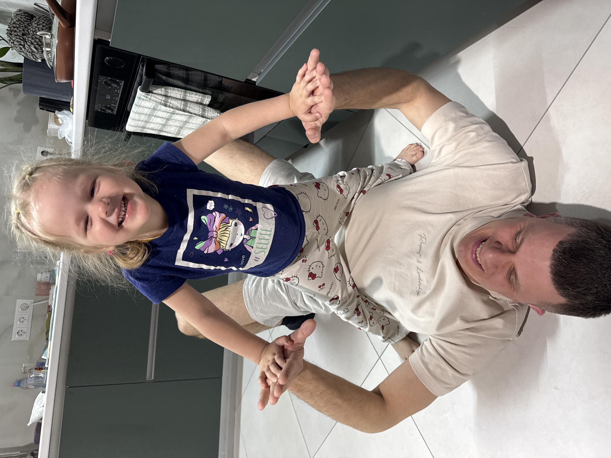 This media file captures a joyful moment between a father and his young daughter, both smiling broadly while lying on a light-colored tiled floor, likely in a kitchen or dining area. The image is rotated 90 degrees clockwise.
The adult male, presumably the father, is on his back, wearing a light beige t-shirt with "Respect & Leadership" printed subtly on the chest, and light gray shorts. He has short dark hair and is looking upwards with a wide smile, his eyes partially closed from happiness. His right arm is extended, holding his daughter's hands.
The young girl, with blonde hair in two small pigtails secured with yellow ties, is propped up on her father's bent left leg. She is facing the camera, her face alight with a huge, open-mouthed smile, eyes closed in delight. She wears a navy blue t-shirt with a colorful cartoon graphic that includes the words "LOVE" and "#SELFIE". Her light gray pajama pants are patterned with "Hello Kitty" characters and text. She holds her father's hands, and her small, bare feet are visible.
In the background, typical kitchen elements are visible: a white countertop, dark green/grey cabinets, a black oven or stove with knobs and a digital display, and plaid towels hanging nearby. Other items include a pot, a plant, a power outlet with three sockets, and various bottles and a plastic bag on the countertop. The scene conveys a strong sense of happiness, playfulness, and family bonding.
