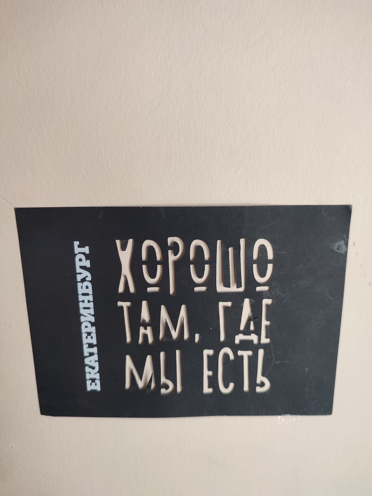 A close-up, eye-level shot displays a black rectangular metal sign with white cut-out letters against a textured light beige wall. The sign is positioned horizontally, slightly angled with the left side lower than the right. The left side of the sign features vertical text that reads "ЕКАТЕРИНБУРГ", with the letters oriented upwards. To the right of this, the primary text is arranged in three lines. The top line says "ХОРОШО", the second line reads "ТАМ, ГДЕ", and the bottom line states "МЫ ЕСТЬ". All text is in uppercase Cyrillic letters. The lighting is even, and there are no shadows cast by the sign on the wall. The image does not provide any information about Kadıköy, Turkey.
