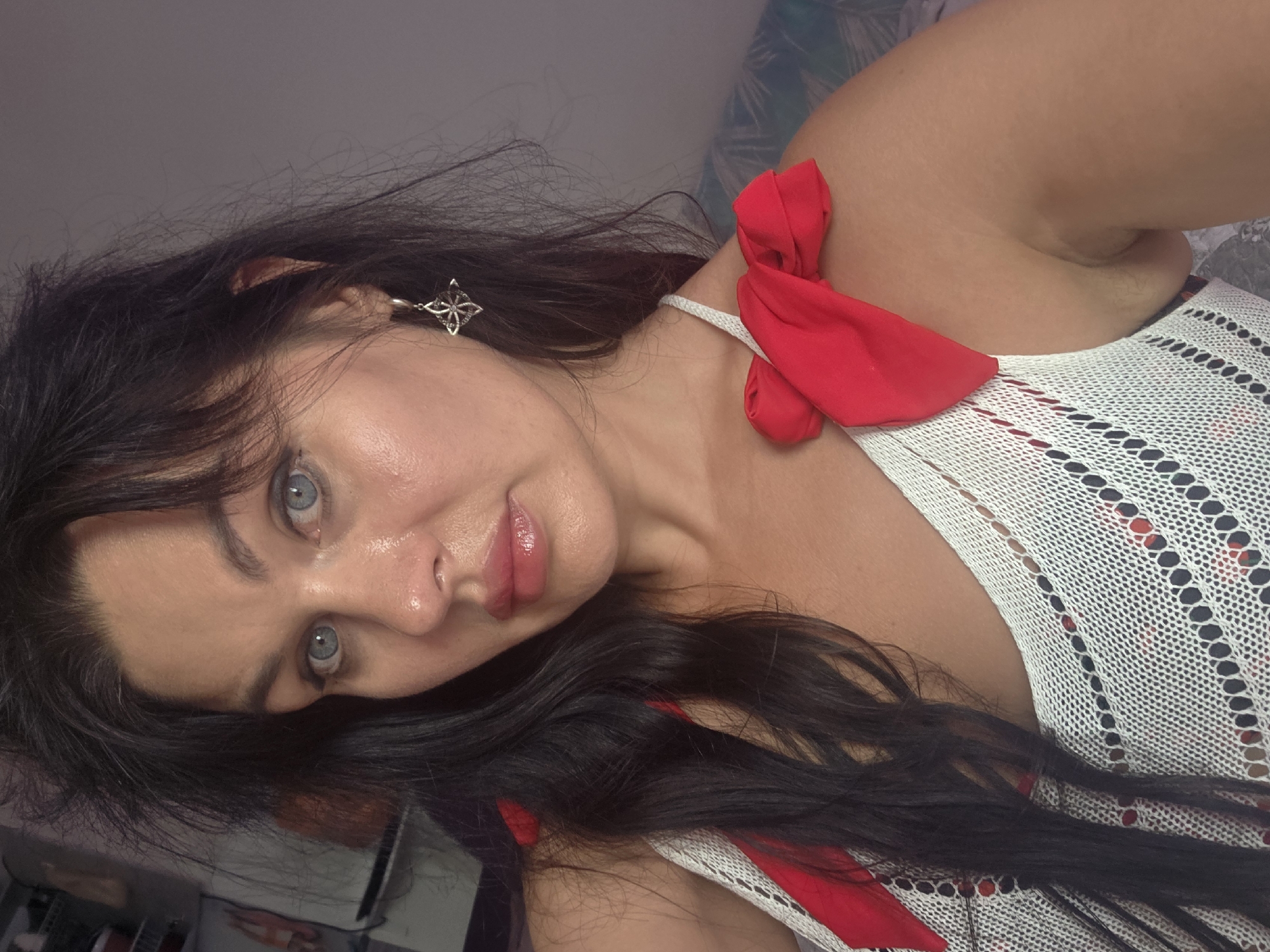 A woman with long, dark hair and blue eyes is shown in a close-up selfie. She is wearing a white top with red ribbon details on the shoulders. She is also wearing a silver earring with a star design. The background is a plain wall. The image does not contain any specific visual cues that would allow for the determination of its location in the city of Sochi, Russia.
