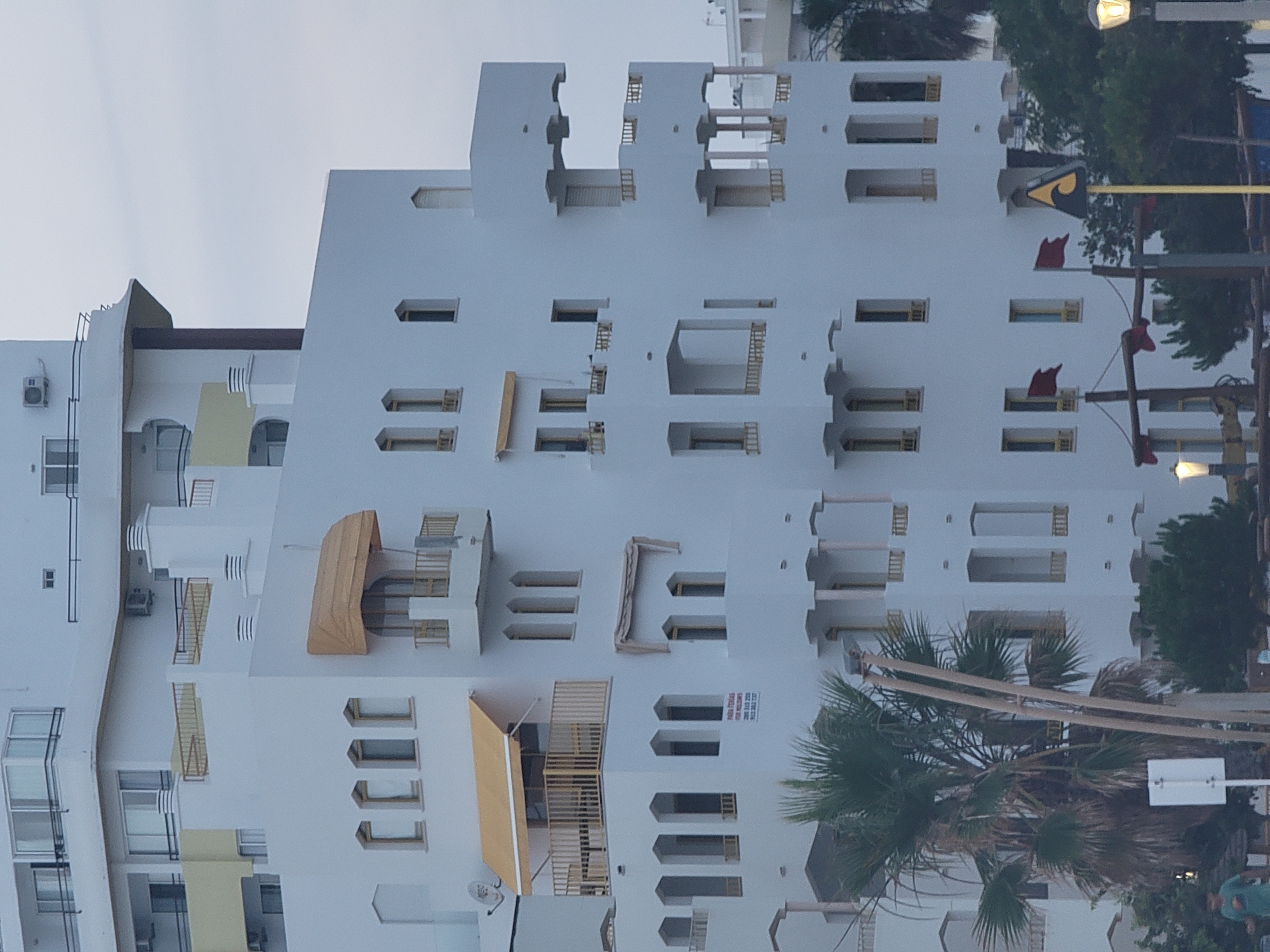 This is a vertical, eye-level, full-shot photograph of a multi-story white building, likely a hotel or apartment complex, in Quarteira, Portugal. The architecture features several balconies with light-colored railings, arched windows with yellow accents, and multiple awnings, some of which are yellow. There are also air conditioning units visible on some of the upper floors.
In the foreground, a tall palm tree with fronds is on the left side of the image, partially obscuring the building. To the right of the palm tree, there is a white directional sign on a pole. Further to the right, a section of a wooden structure, possibly part of a playground or decorative element, is visible with red flags attached. A streetlamp is also present on the far right.
In the middle ground, a person in a blue shirt and shorts is walking from left to right. The sky is overcast and a pale gray. The overall impression is a daytime scene on a cloudy day in a coastal town.