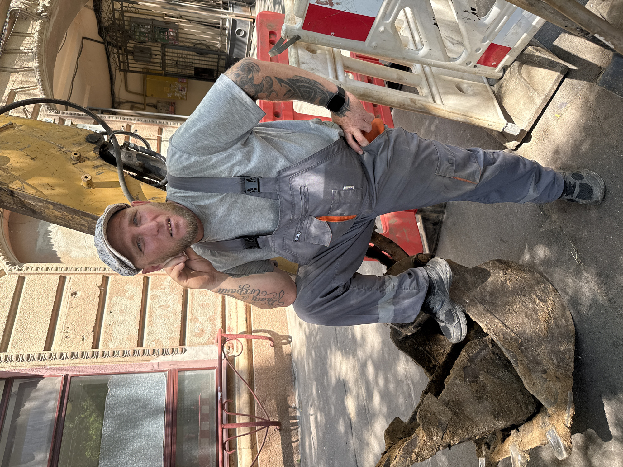 The image shows a man standing next to an excavator. He is wearing grey overalls and a grey t-shirt, and a cap. He has tattoos on his arms and left leg. The man is looking at the camera and smiling. He appears to be working, as he is dressed in work clothes and is near construction equipment.
The scene appears to be outdoors, possibly on a street or construction site. The excavator is yellow and appears to be partially buried in dirt or mud. There are orange and white safety barriers in the background. The building behind the man has decorative architectural features and a red-trimmed window.
The lighting suggests it is daytime, with shadows cast on the ground. The man's expression is friendly and relaxed, indicating a positive mood despite the likely physically demanding nature of his work. There is no visible text in the image, although there is a logo on the overalls which is too small to be legible.