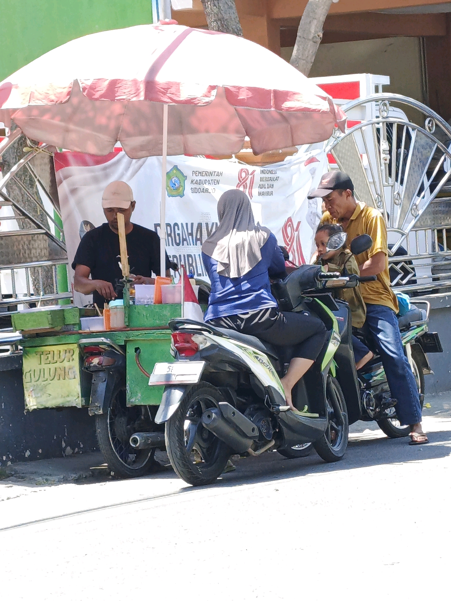 The image depicts an outdoor scene, likely on a street in Ketegan, Indonesia, with a food vendor operating from a mobile cart attached to a motorcycle. The vendor, a man wearing a black t-shirt and a light-colored cap, is preparing food. He is positioned behind a green cart displaying various containers and utensils. A banner with Indonesian text and government seals is visible behind him.
Two people are seated on a scooter parked next to the vendor's cart. A woman wearing a grey hijab and blue shirt is on the scooter, and a man in a yellow shirt and blue jeans is behind her, holding a child who is also on the scooter. The scooter is adorned with green and white accents.
Another motorcycle is parked to the left of the vendor's cart. The environment appears to be a public space with a tree and a building in the background. The overall lighting suggests a sunny day. The sign on the green cart reads "TELUR GULUNG," which translates to "rolled egg," indicating the food being sold.