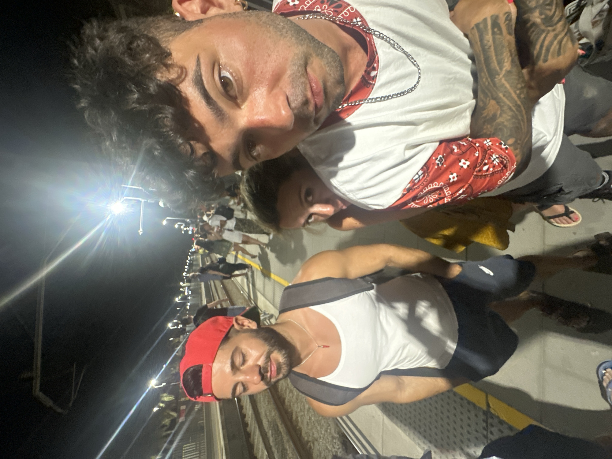 The image depicts a group of people on a train platform at night, likely in El Masnou, Spain, given the context of the provided crops which hint at Spanish signage. The central focus is a man in the foreground, wearing a white tank top, dark shorts, and a red baseball cap. He has a backpack on and is wearing a necklace with a red pendant. To his left, another man with a red cap and white tank top is visible, looking down. Behind the man in the foreground, a woman with light brown hair is peeking from behind him, looking directly at the camera. In the background, other people are standing on the platform, some with their backs to the camera, and train tracks are visible. The platform is illuminated by artificial lights, creating strong highlights and shadows. The overall scene suggests a casual gathering or travel at a public transportation hub during nighttime hours.