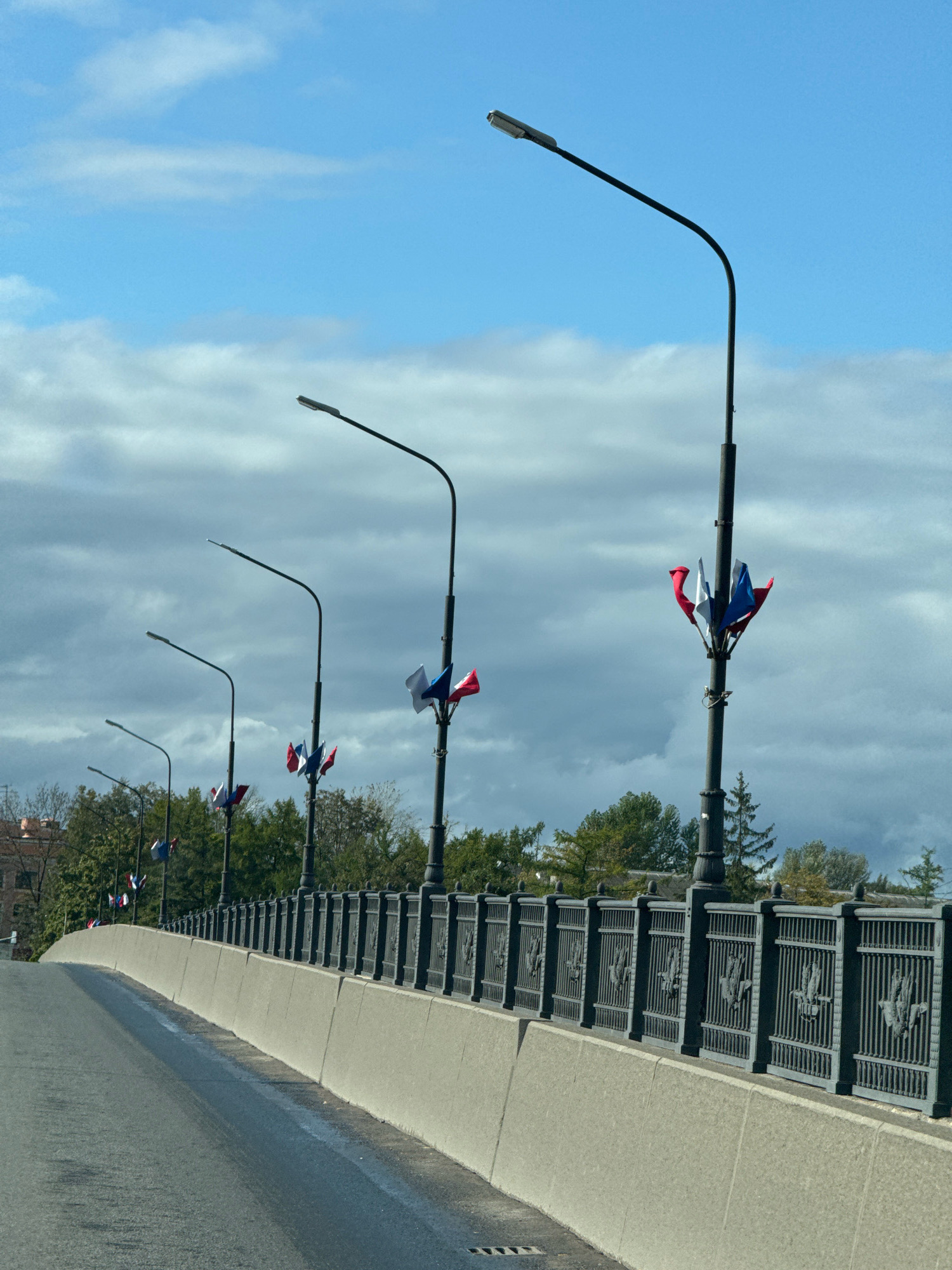 The image shows a view from a car driving across a bridge in Veliky Novgorod, Russia. The bridge has a concrete retaining wall and a decorative metal fence along the side. Several lampposts with curved arms extend over the bridge, and some are adorned with small red, white, and blue flags, possibly a national flag. Trees and buildings are visible in the background, with a cloudy sky overhead. The road surface appears to be asphalt and is wet in some areas.