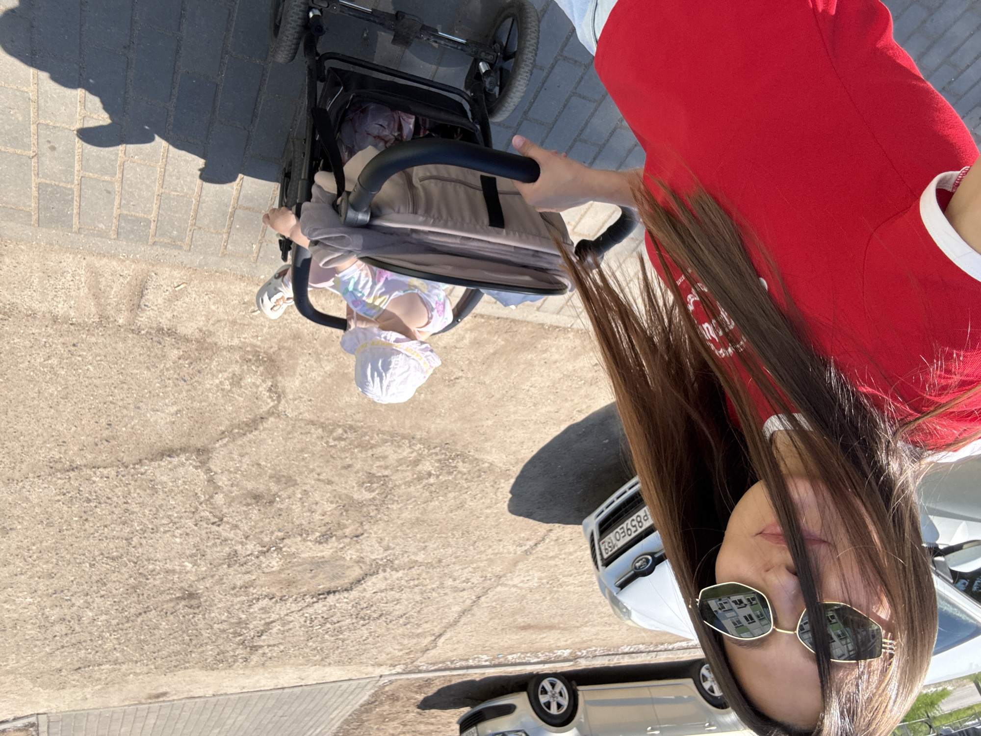 A young woman with long brown hair, wearing a red t-shirt with white trim and sunglasses, is taking a selfie outdoors. She is pushing a baby in a stroller. The baby is wearing a white sun hat and a colorful shirt. In the background, two cars are parked on a dirt and asphalt road. One car, a silver sedan, is in the upper background, and a white Ford is in the middle ground with a Russian license plate that reads "P859EO 159". The scene appears to be in a residential area in Chaikovsky, Russia, given the license plate. The ground is a mix of dirt, gravel, and paving stones. The lighting suggests it is a sunny day.