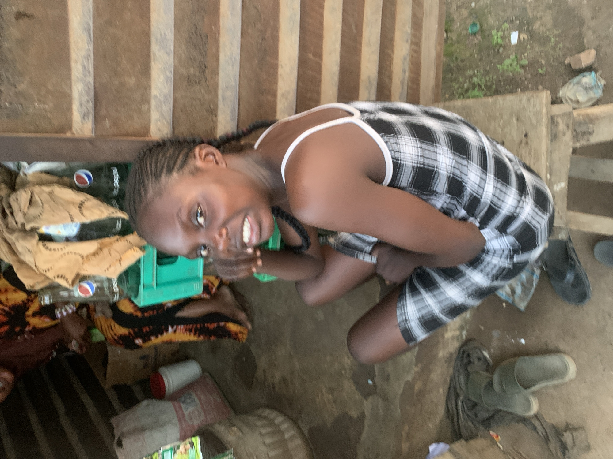 The image captures a young girl with braided hair, wearing a black and white checkered dress, sitting on a wooden plank outdoors. She is looking towards the camera with an open mouth, possibly speaking or reacting. Behind her, to the left, is an area with various items including bags, bottles of beverages (including Pepsi), and a green crate. A woman in a vibrantly colored dress with yellow, orange, and black patterns is partially visible in the background, seated on the ground. The environment appears to be a street-side stall or shop, characterized by rough wooden walls and a dirt ground. The presence of local products and the setting suggest a scene likely found in a bustling area of Shagamu, Nigeria.