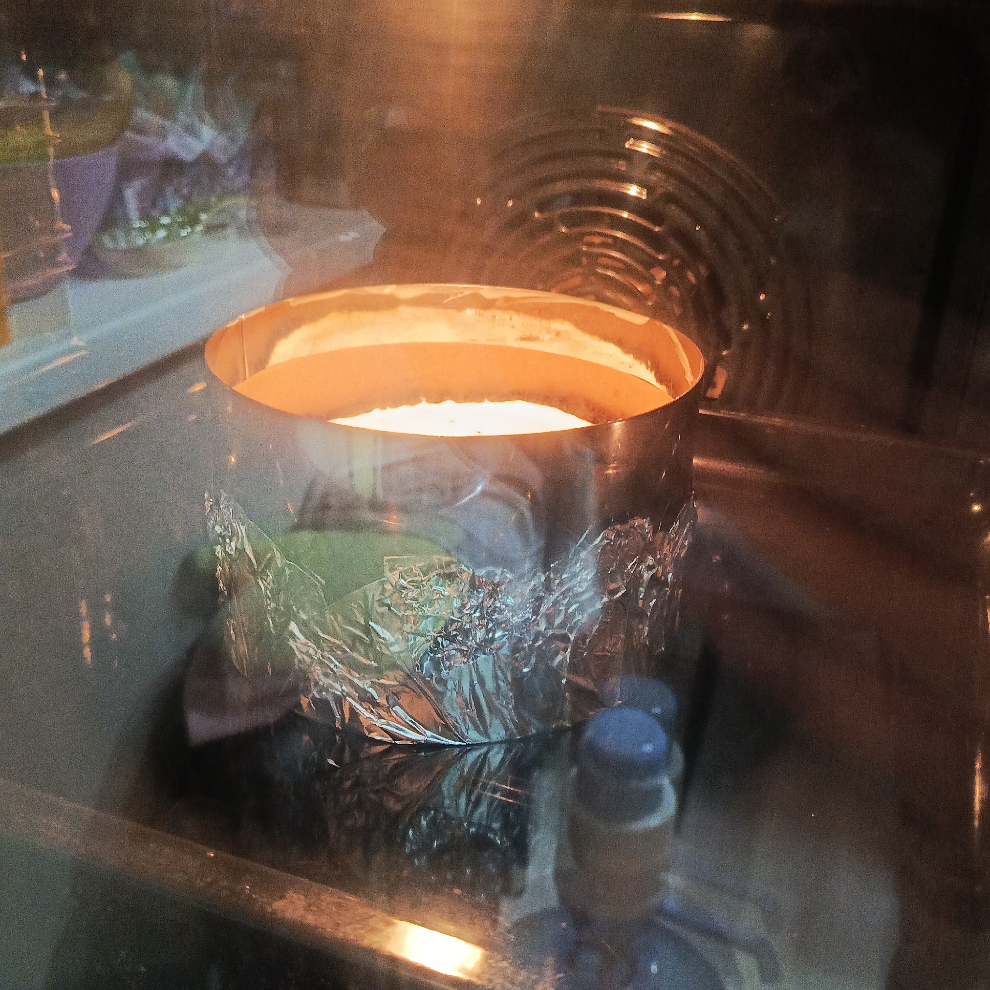 Inside an oven located in the city of Марковский, Russia, a circular metal baking pan contains a light-colored substance, likely a batter or dessert, undergoing a baking process. The lower exterior of the pan is wrapped in crinkled aluminum foil. The oven's interior is brightly illuminated by an intense orange light source, casting a warm glow over the substance's surface and the internal reflective surfaces. A coiled heating element is visible in the upper right background. Reflections on the oven door glass distort views of the surrounding environment, showing blurred colors and indistinct shapes. A partial view of a blue-capped object is present in the lower right foreground, visible through or reflected on the oven door. No individuals are present.