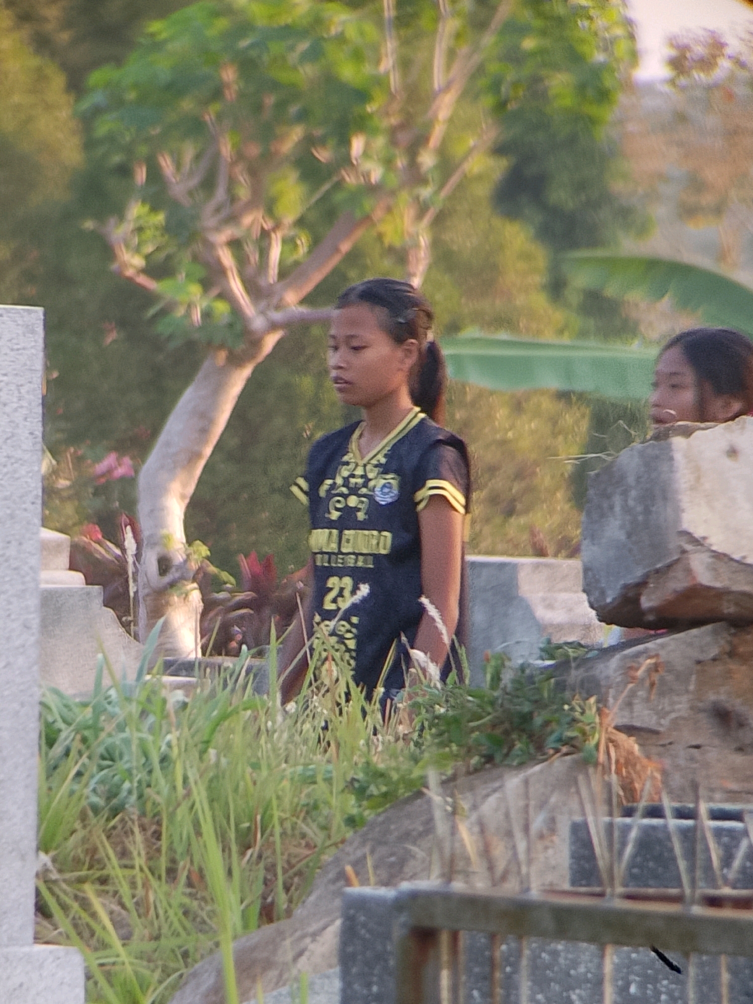 The image shows two young women in a graveyard. The woman on the left is wearing a dark shirt with yellow trim and the number 23 on it. She has her hair pulled back in a ponytail. The woman on the right is partially obscured by a concrete structure and is looking to the right. The background features trees and greenery. The overall scene suggests a sunny day. The provided crops indicate the location may be in Jember, Indonesia.