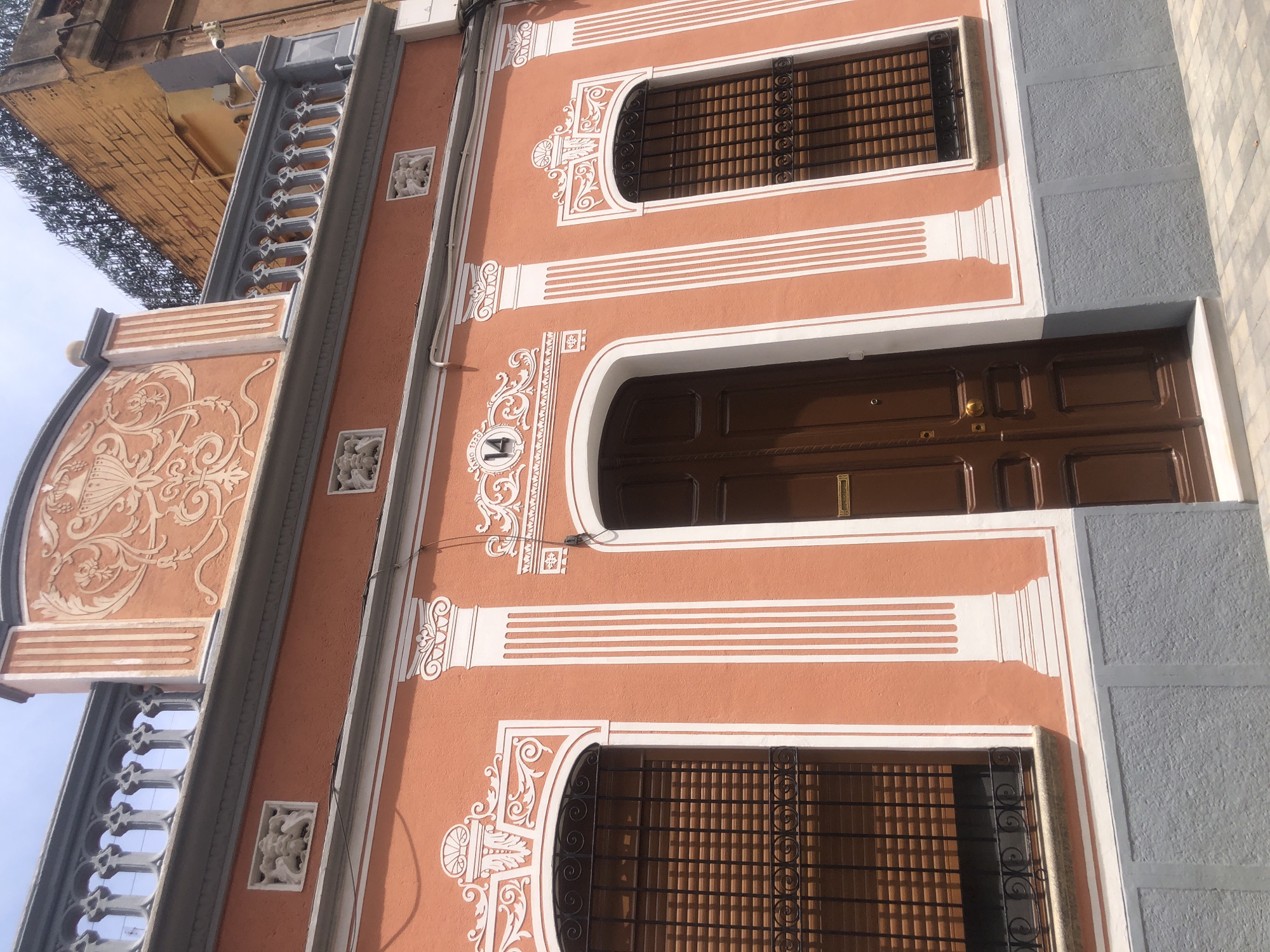 The image shows the facade of a building with an orange-colored stucco exterior and gray stucco on the lower portion. The building features decorative white trim around the windows, door, and along the roofline. There are two windows with black wrought iron grilles and wooden blinds. A brown wooden double door is centrally located, flanked by decorative columns. Above the door, the number "14" is visible within a decorative white frame. The roofline is adorned with a balustrade and ornate stucco designs. The sky is visible above. The scene appears to be an exterior shot of a residential building.
