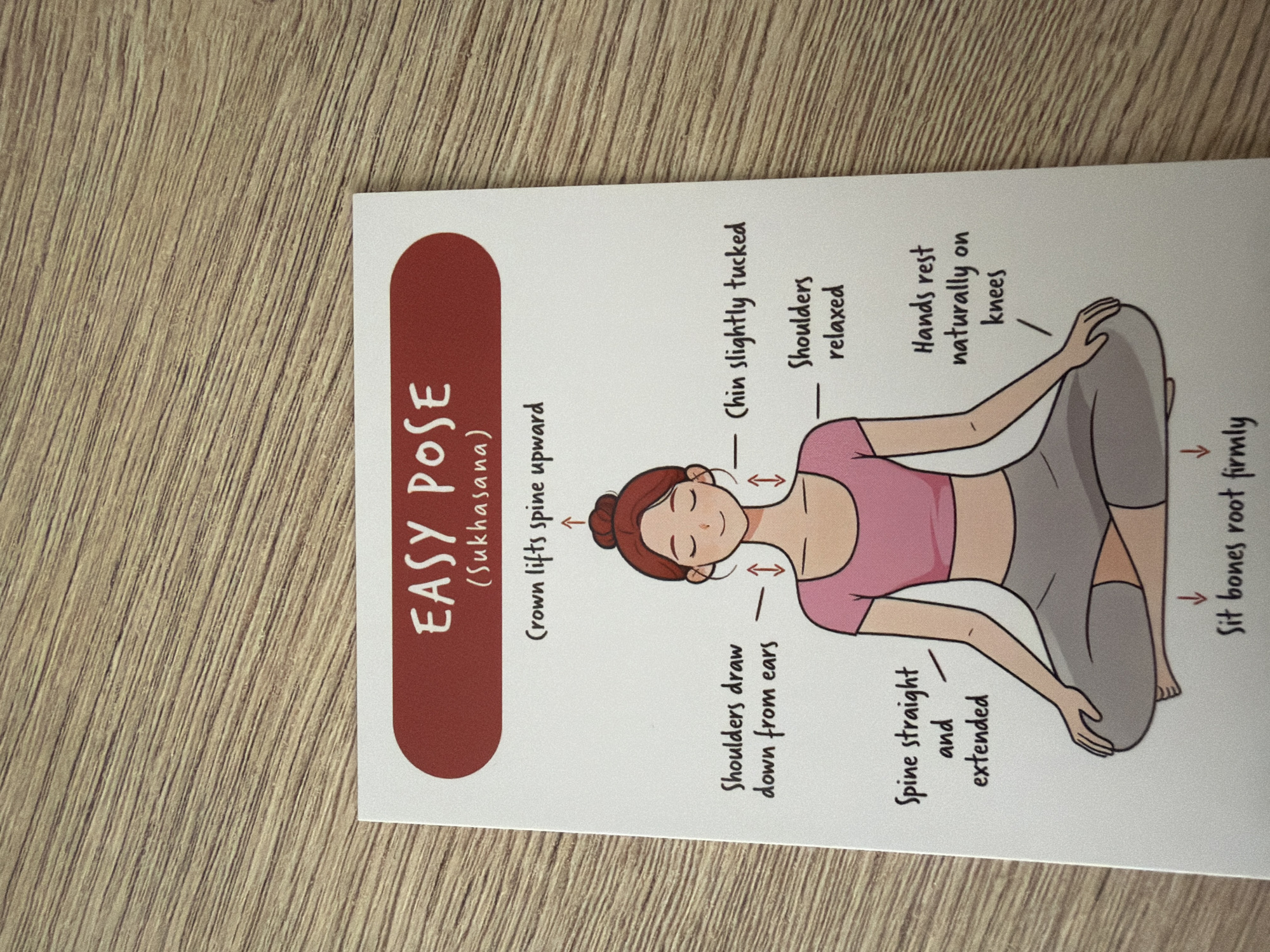 A postcard depicting the easy pose (Sukhasana) in yoga is shown on a wooden surface. The postcard has a maroon header with white text reading "EASY POSE (Sukhasana)". Below the header, an illustration of a woman in a yoga pose is shown with instructional text. The woman is sitting cross-legged with her eyes closed and a serene expression. Arrows and text point to different parts of her body, indicating proper posture: "Crown lifts spine upward", "Shoulders draw down from ears", "Chin slightly tucked", "Shoulders relaxed", "Spine straight and extended", "Hands rest naturally on knees", and "Sit bones root firmly". The postcard is angled on a light brown, textured wooden surface, likely a table. There is no information about the city of Pazardzhik, Bulgaria, in this image.