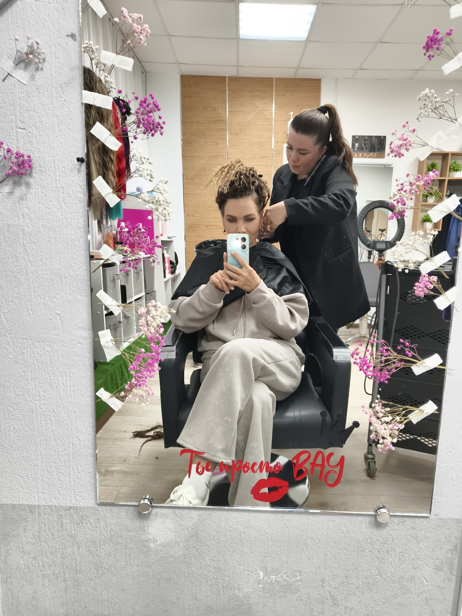 The image shows a mirror selfie taken by a client in a hair salon or studio. In the foreground, a woman with light-colored, dreadlock-style hair is seated in a black salon chair, wearing a light grey tracksuit and a black protective cape. She is holding a light blue/green smartphone to take the picture, with her face partially visible and an engaged expression.
Behind her, a female stylist, dressed in a black jacket with her dark hair tied in a high ponytail, is actively working on the client's hair. She is focused on her task, using tools near the client's head.
The large mirror, which serves as the canvas for the selfie, is decorated with small sprigs of pink and white baby's breath flowers secured with white tape. Various hair extensions or wigs in different colors (browns, reds, teal) are also displayed vertically on the mirror. The reflection reveals parts of the salon's interior, including shelves stocked with hair products, a black rolling cart, and a ring light stand. The background features a patterned wooden wall panel and a white textured wall, along with white ceiling tiles and fluorescent lighting.
On the lower right corner of the mirror, red text is visible. It reads "Ты просто BAY" with a prominent red lipstick kiss mark graphic replacing part of the word "просто". The overall lighting suggests it is daytime.