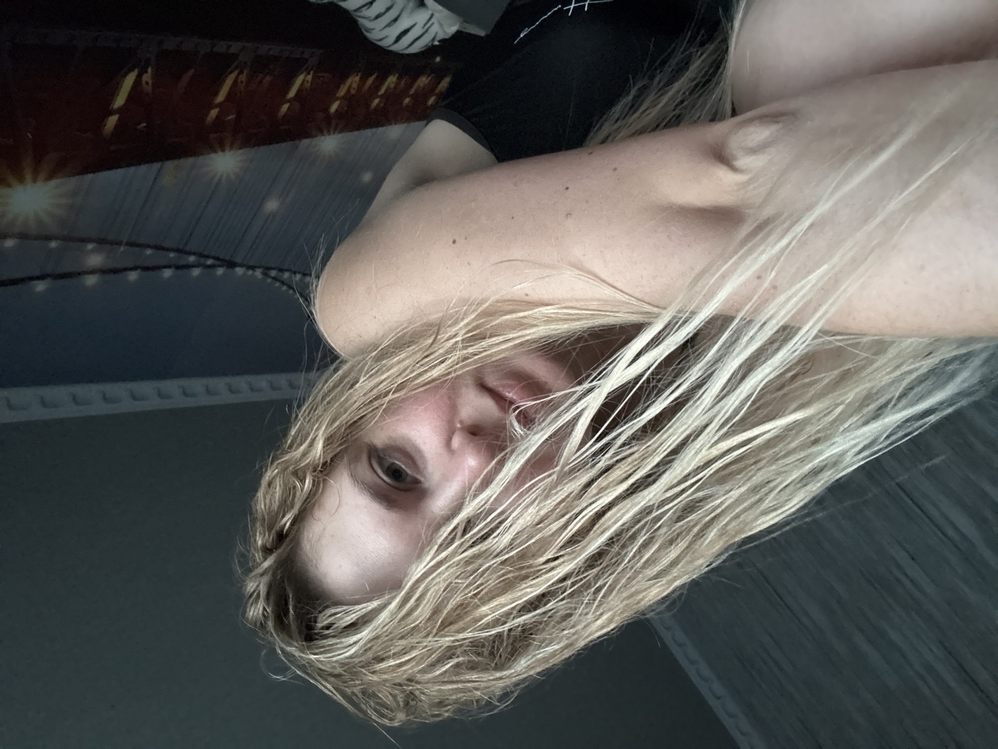 A young woman with blonde, wet hair is captured in a close-up, low-angle shot. She is leaning on her arm, with her hair partially obscuring her face. Her gaze is directed towards the camera with a slightly melancholic expression. The background appears to be a dimly lit room with a patterned wall and a decorative element that resembles a bridge with lights. The overall lighting is soft, highlighting the texture of her hair and skin.