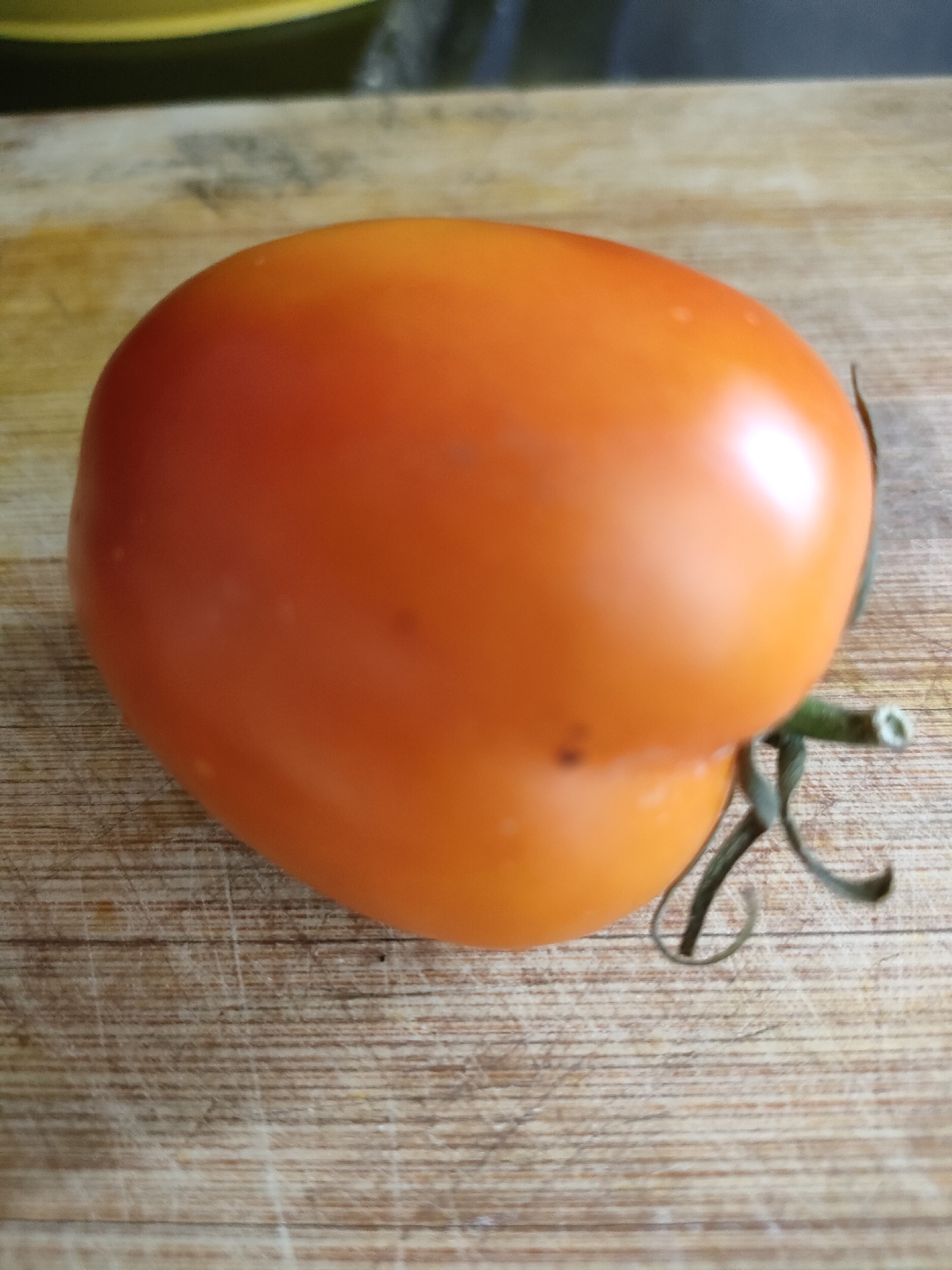 A single, ripe, orange tomato rests on a wooden cutting board. The tomato is slightly oblong and has a smooth, shiny skin. A small green stem with three tendrils is attached to the top of the tomato. The cutting board has a light brown, wood grain pattern with visible scratches and marks from use. The background is blurred but appears to be a dark surface with a hint of yellow. The scene is lit from the top, casting a subtle shadow. The location is Unknown, Indonesia.