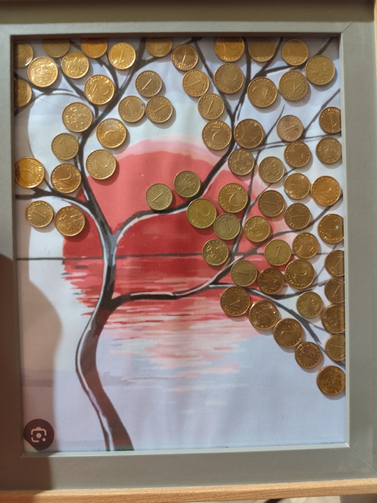 The image shows a framed artwork featuring a tree with coins as leaves and branches against a backdrop of a stylized sun and water. The coins are gold-colored and appear to be of different denominations, some with visible inscriptions. A small camera icon is present in the bottom left corner of the frame. The image does not contain any specific geographical markers or elements that would allow for identification of its location in the city of Montana, Bulgaria.