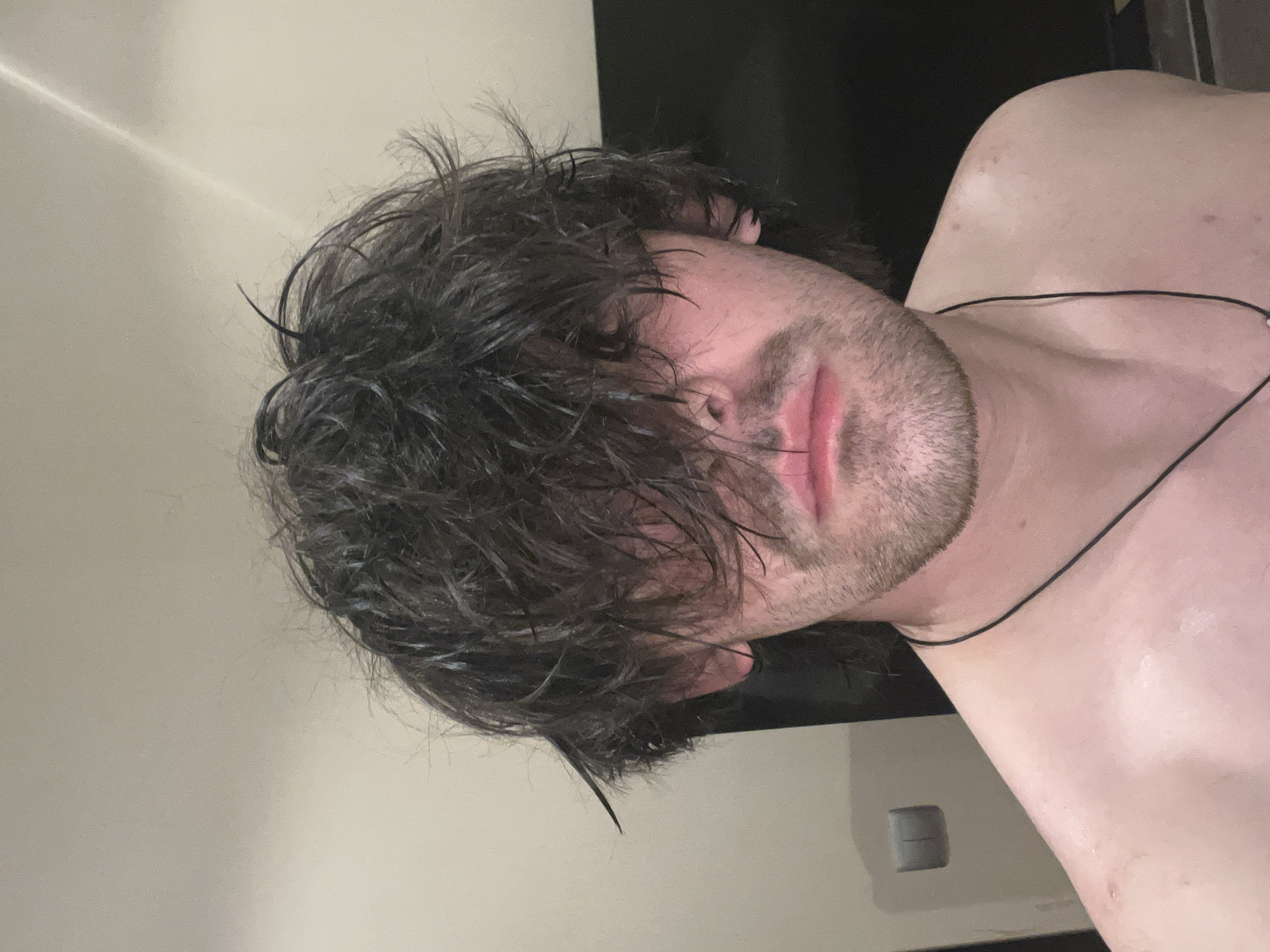 A close-up, portrait-oriented shot features a fair-skinned male with wet, dark hair partially obscuring his face. The hair is tousled and covers much of his forehead and part of his eyes. He has a light beard and mustache. He is wearing a thin, black necklace. The background consists of a light-colored wall on the left, transitioning to a dark-colored surface on the upper right. The scene also includes what appears to be a light switch or other wall-mounted device lower right. The setting is not recognizably in Ореховка, Russia. The photo does not contain contextual cues indicating location.