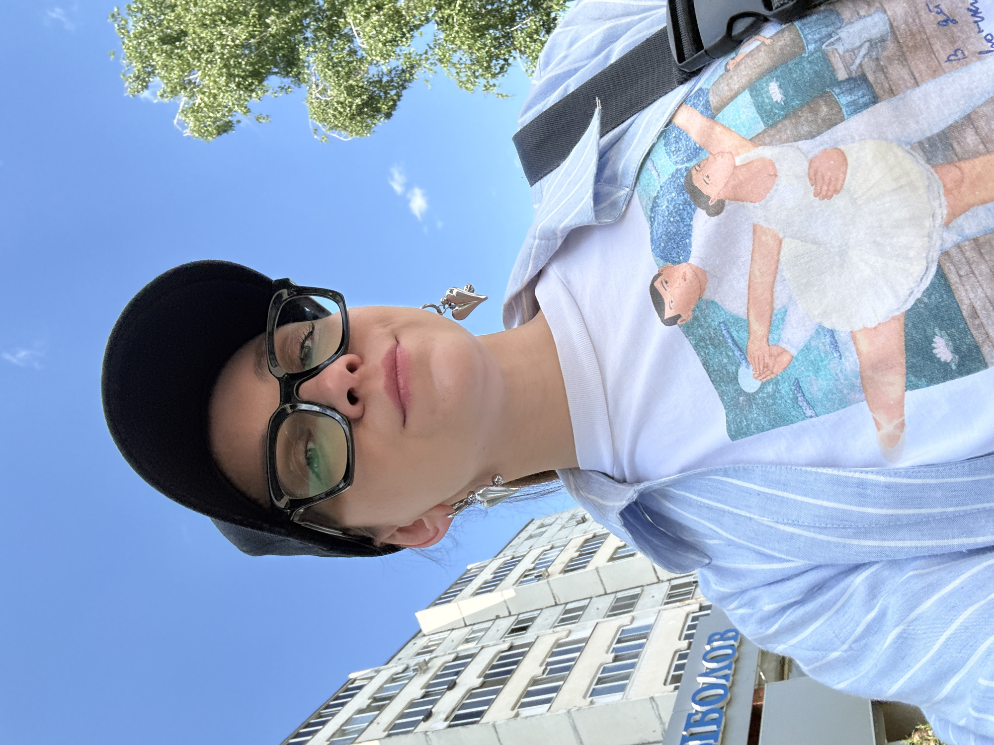 A low-angle photograph shows a person wearing glasses and a black cap looking down at the camera, with a building and blue sky in the background. The person wears a white t-shirt with a design of a dancing couple and a light blue striped shirt. The building is tall with multiple windows. Blue letters spelling out "ПИБОЛОВ" are visible on the building. The location appears to be in Kazan, Russia, based on the Cyrillic letters on the building. A tree is partially visible at the top of the frame. The person also wears dangle earrings. A black strap is draped across the person's shoulder.