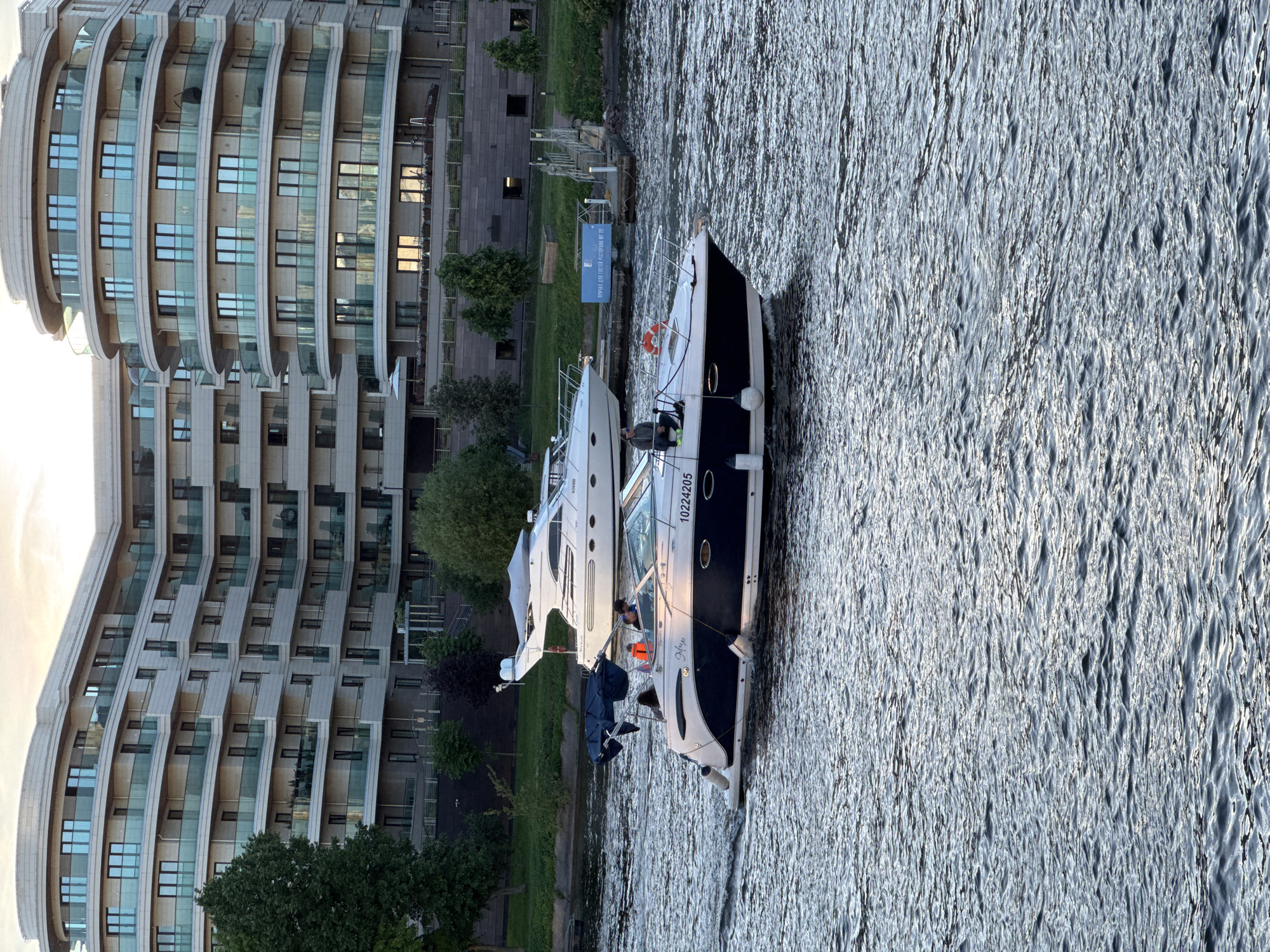 The image captures a scenic view of the Neva River in Saint Petersburg, Russia, with modern residential buildings in the background. A sleek, dark blue and white motor yacht, identified by the number "10224205" and the name "Mona" on its hull, is the primary focus. It's moving from left to right, creating ripples on the water's surface. Several people are visible on board: one individual is steering, another is seated near the bow, and two others are in the stern, one wearing an orange life vest. Behind this yacht, a larger white motor yacht is docked or anchored, also with people visible on its deck. The buildings in the background are contemporary, multi-story structures with curved balconies and large windows, reflecting the soft light of what appears to be late afternoon or early evening. Greenery, including trees and grass, lines the riverbank in front of the buildings. A small pedestrian pier with a gangway extends into the water. A blue sign with Russian text is visible on the pier. The overall impression is one of urban riverside leisure.