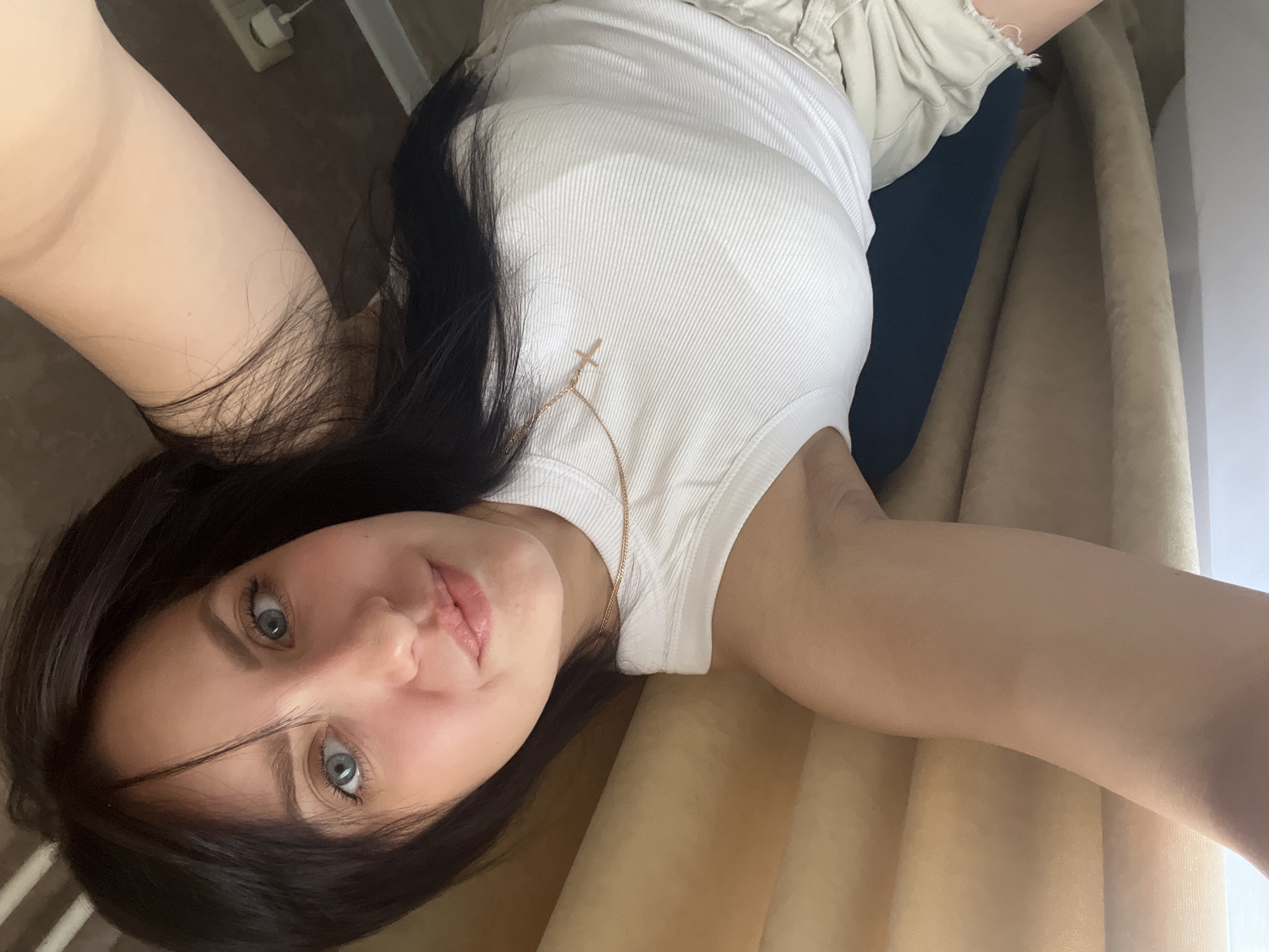 A young woman with dark hair and light blue eyes is captured in a close-up selfie, lying down with her head tilted to the side. She is wearing a white, ribbed tank top and light-colored shorts. A delicate gold chain with a cross pendant rests on her chest. Her right arm is visible, extended upwards. The background appears to be an indoor setting, possibly a bedroom or living area, with textured brown walls and a beige curtain. A white electrical outlet with a plug is visible on the wall in the upper right corner. The image does not contain any specific visual cues that would allow for its location to be identified within Марковский, Russia.