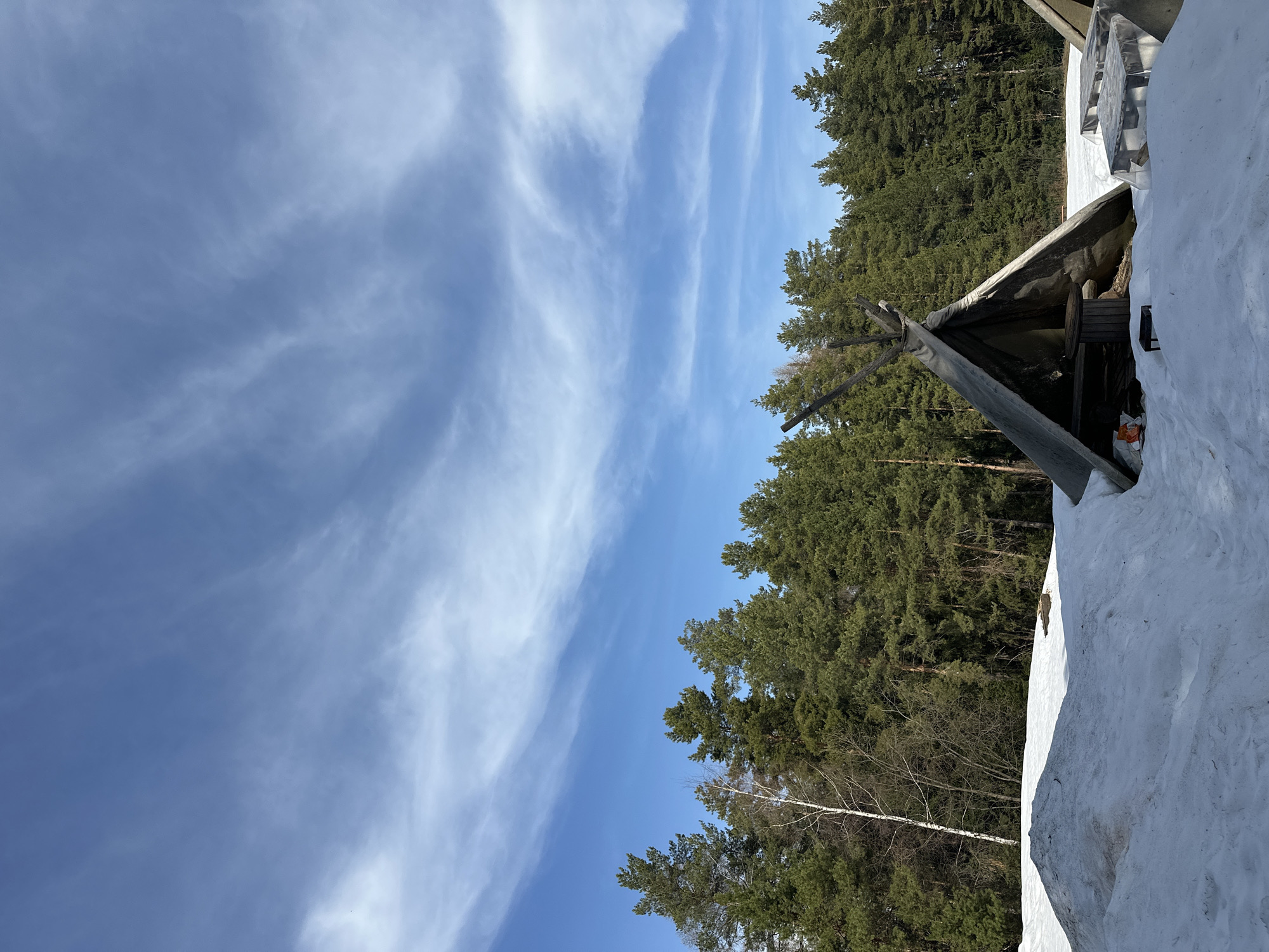 The image depicts a wintry scene in Прикамский, Russia. The sky is bright blue with streaks of cirrus clouds. A dense forest of evergreen trees stretches along the right side of the frame. A tipi-like structure, possibly made of wood and canvas, is partially visible on the right, covered in snow. The ground is also covered in snow, suggesting a significant snowfall. There are no people visible in the frame. The overall impression is of a rural or wilderness setting on a clear winter day in Прикамский.