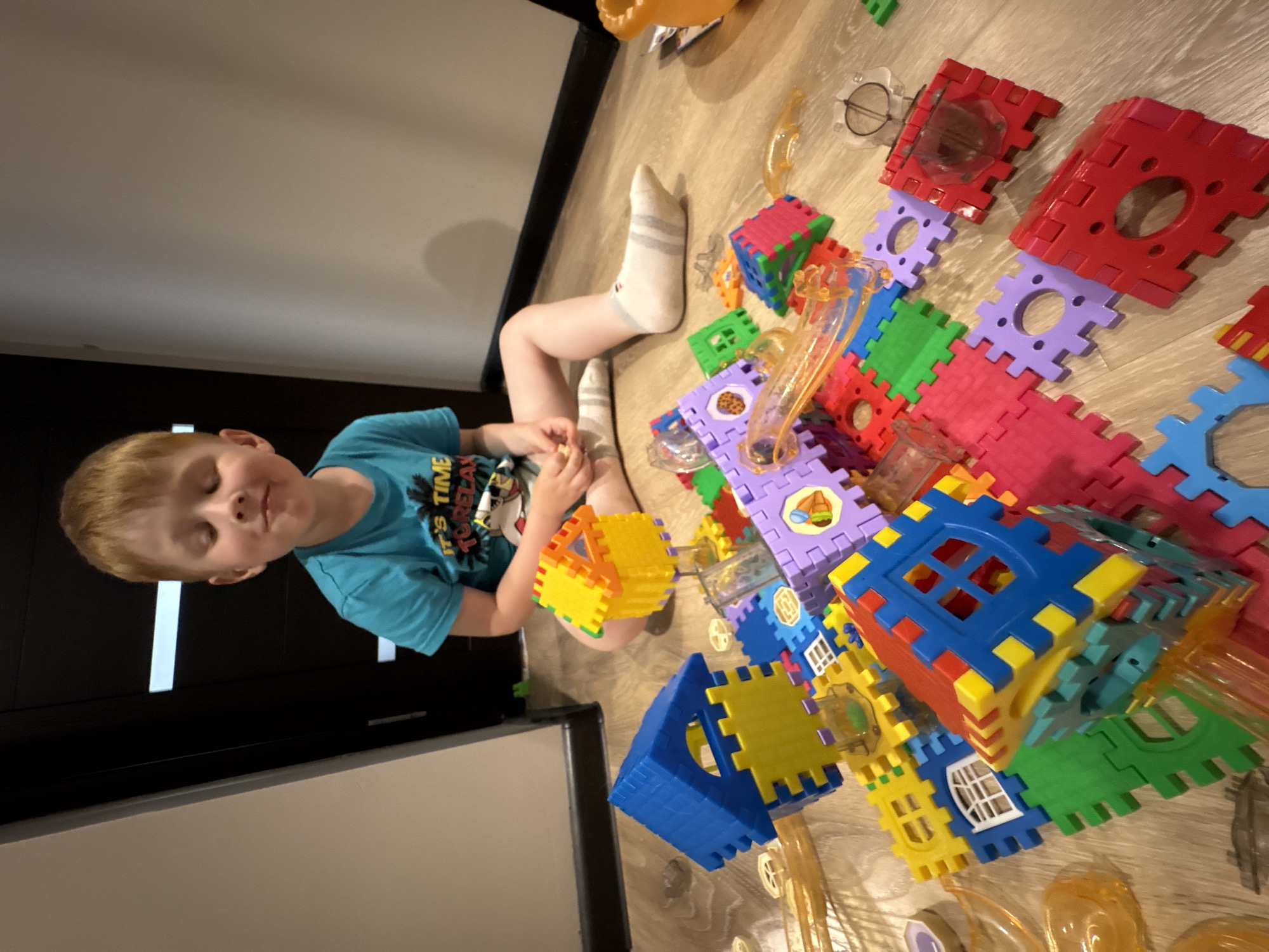 A young boy with blond hair and fair skin, wearing a turquoise t-shirt and gray socks, sits on a wooden floor surrounded by colorful building blocks. The blocks are made of plastic and come in various shapes and sizes, including square, rectangular, and triangular pieces, in colors such as red, blue, green, yellow, and purple. Some of the blocks are assembled into structures resembling houses. He appears to be looking down at the toys, with his eyes closed and a slight smile on his face, suggesting he is engaged in play. The scene takes place indoors, likely in a home, given the wooden floor and the presence of children's toys. The lighting suggests it could be daytime. The boy's t-shirt has text on it that is partially visible, including "IT'S TIME TO RELAX" and a graphic of a palm tree. The setting is Izhevsk, Russia.
