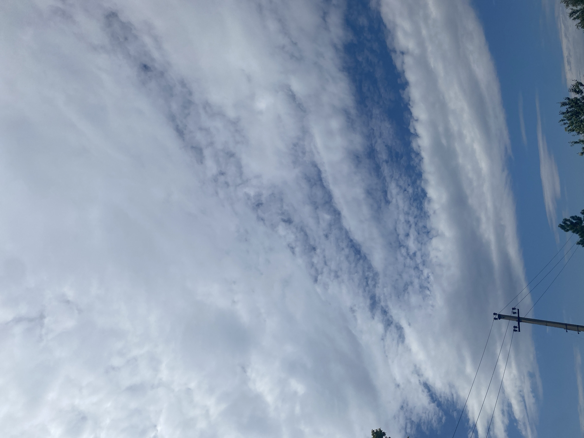 The image captures a bright, partly cloudy sky over Oktyabrsk, Russia. Wispy white clouds streak across a vibrant blue sky, with denser cloud formations lower in the frame. A single utility pole, with wires extending to the left, stands in the lower left quadrant of the image. The bottom edge of the image shows the tops of green trees.