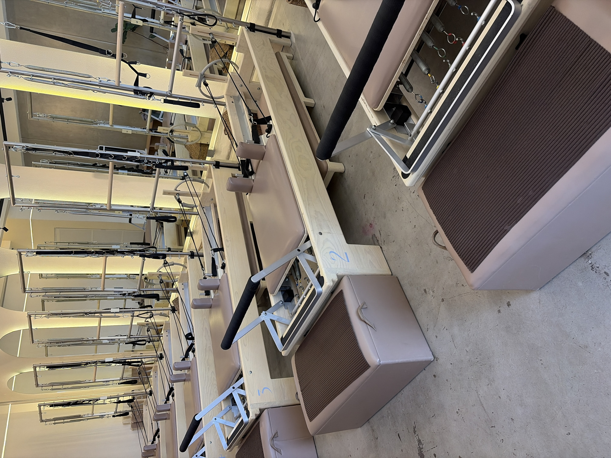 The image captures a modern Pilates studio, devoid of people, showcasing multiple pieces of exercise equipment. The primary subjects are several Pilates reformers, arranged in rows and extending from the foreground into the midground. These reformers feature light-colored wooden frames, padded beige carriages, and various metallic components including springs, pulleys, and foot bars. Black straps and handles are attached to the machines, and black padded bars are also visible.
Interspersed with the reformers are light beige boxes with dark brown ribbed surfaces, likely Pilates jump boards or exercise boxes, one of which shows a small fabric handle. The floor is a light grey concrete surface with some minor scuffs. In the background, additional Pilates apparatus, possibly Cadillac or Trapeze tables, are visible with their tall metallic frames and suspended elements. The studio walls are light-colored, featuring arched recesses that may be illuminated.
The setting is an indoor studio, brightly lit by what appears to be artificial light. No specific activity is occurring. Visible text includes light blue numbers "2" and "1" (or partially obscured "U"/"V") stenciled on the wooden frames of some reformers, likely indicating station numbers. The image is taken from a slightly elevated, oblique angle and is rotated, presenting the equipment in a diagonal orientation across the frame. The studio appears well-organized and clean.