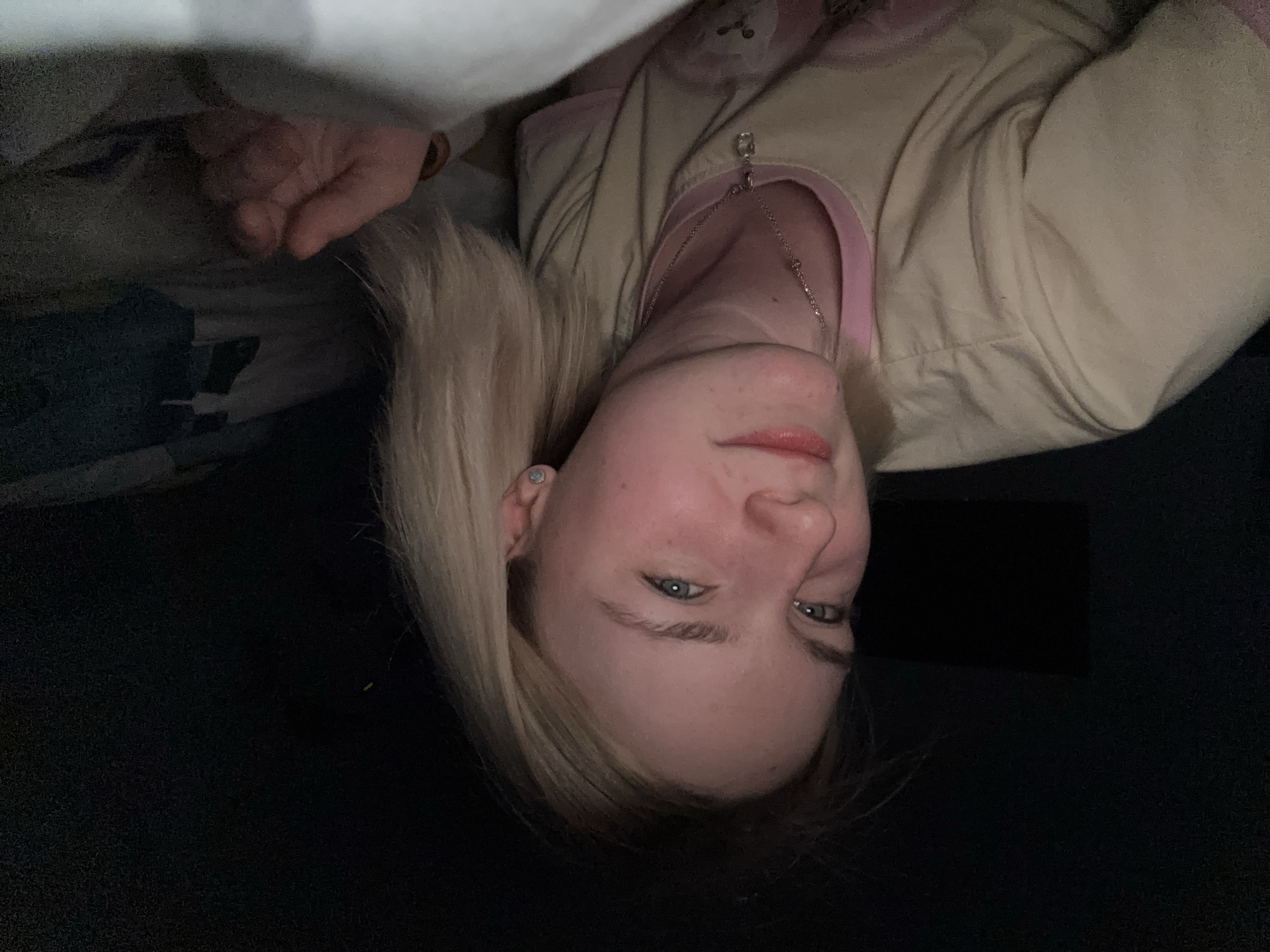 A woman is lying down, possibly in bed, in a dimly lit room. She has blonde hair and is wearing a light-colored pajama top with pink trim and a necklace. Her face is visible, and she appears to be looking at the camera. Part of a hand is visible near the top left corner, possibly holding a blanket. The background is dark and blurry. There is no indication of Vsevolozhsk, Russia.