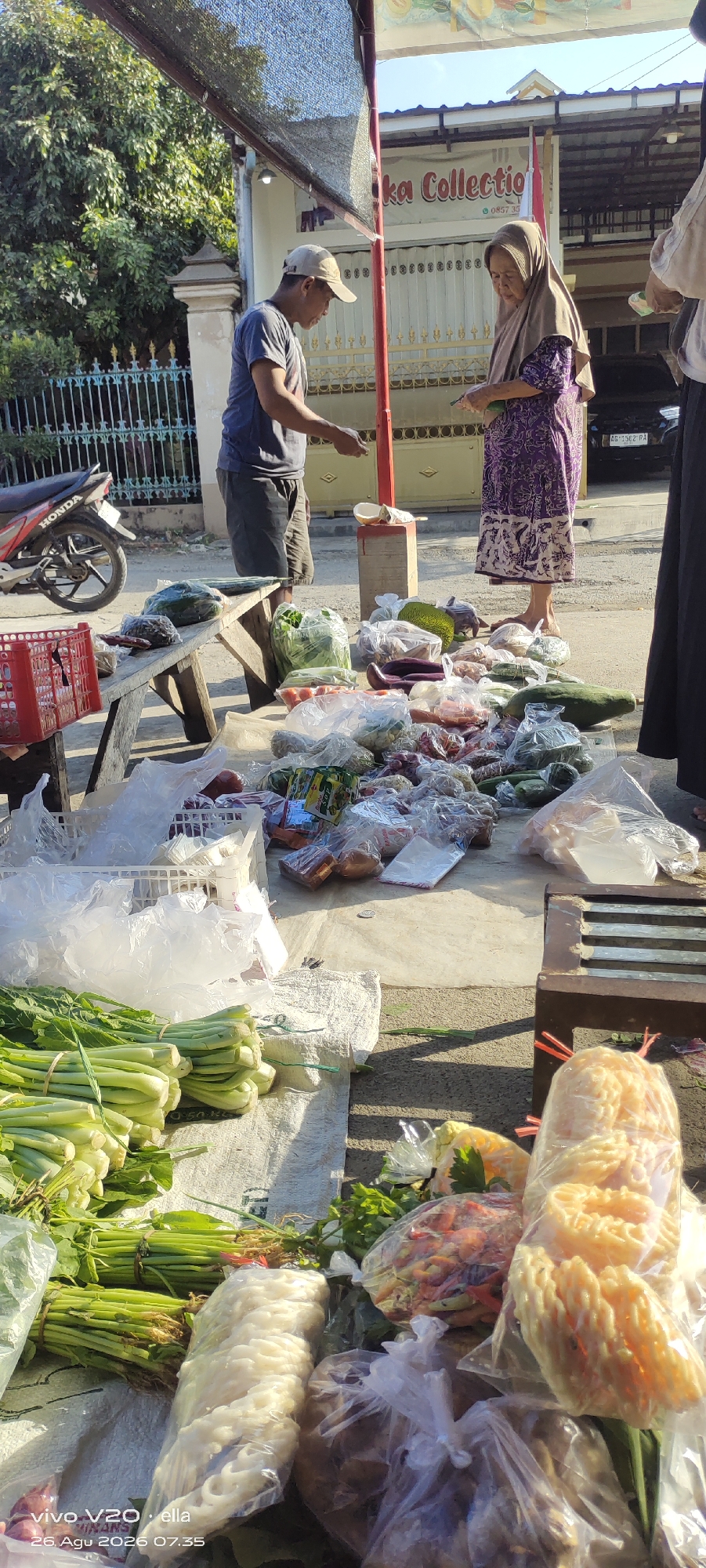 An outdoor marketplace scene is depicted in Simo, Indonesia, under bright daylight. A man wearing a dark t-shirt and a cap stands to the left, reaching out to interact with an elderly woman. The woman, dressed in a purple patterned dress and a light brown headscarf, stands to the right, holding money. Between them, on a wooden bench, are various fresh vegetables and fruits, including leafy greens, cucumbers, tomatoes, and jackfruit, some of which are packaged in plastic. In the foreground, more produce is laid out on a tarp, featuring bundles of green vegetables and various packaged items, including what appear to be crackers and noodles. Behind the people, a building with the text "ka Collectio" is visible, along with a fence and a motorcycle parked on the left. The overall impression is of a typical local market transaction occurring in an urban setting.
