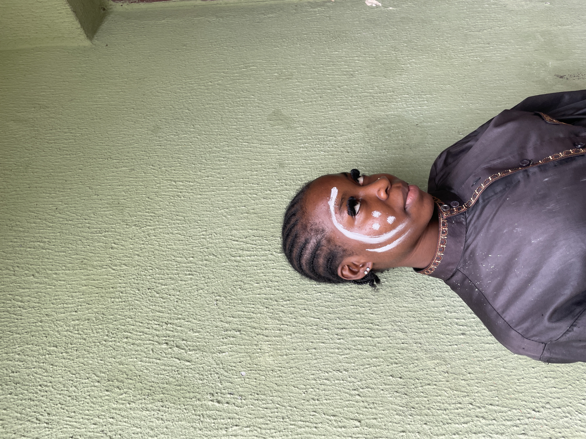 In Ado, Nigeria, a young Black female with braided hair and white face paint is pictured outdoors against a textured, pale green wall. She is looking to her right with a slight smile and her head tilted. The face paint consists of white dots on her cheek and a larger white crescent shape around her eye, with a white stripe above her eyebrow. She is wearing a black, collared shirt with intricate gold trim around the collar and down the front. The wall behind her appears to be roughly plastered and shows some signs of wear. The lighting suggests it is daytime.